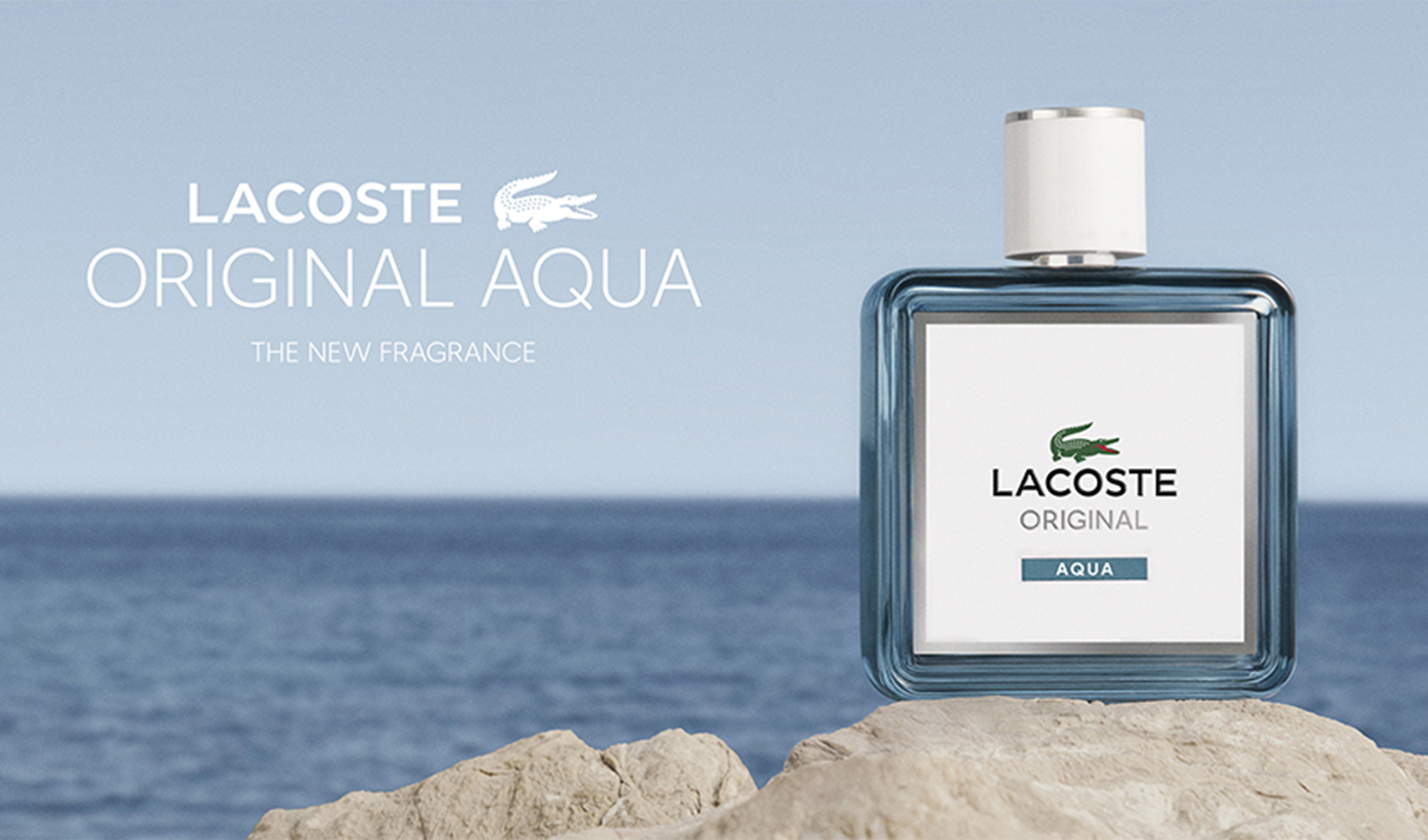 Lacoste Original Aqua parfymeflaske på en stein ved havet, med lyseblå bakgrunn og teksten «Lacoste Original Aqua – The New Fragrance».