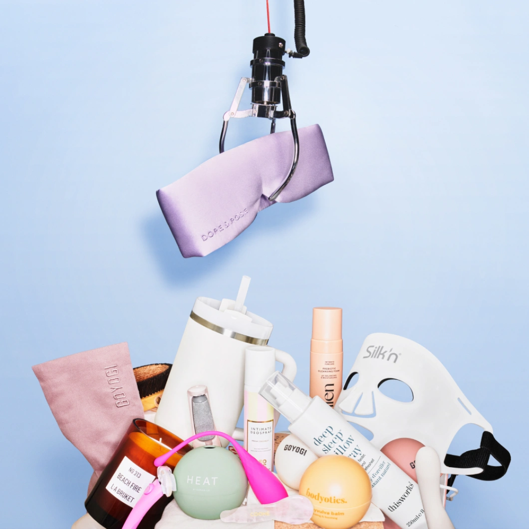 En kloarm holder et lilla silkelignende stoff over ulike wellnessprodukter mot lys blå bakgrunn. Produktene inkluderer Silkn LED-maske, drikkekopp, kroppspleie og wellness-tilbehør.