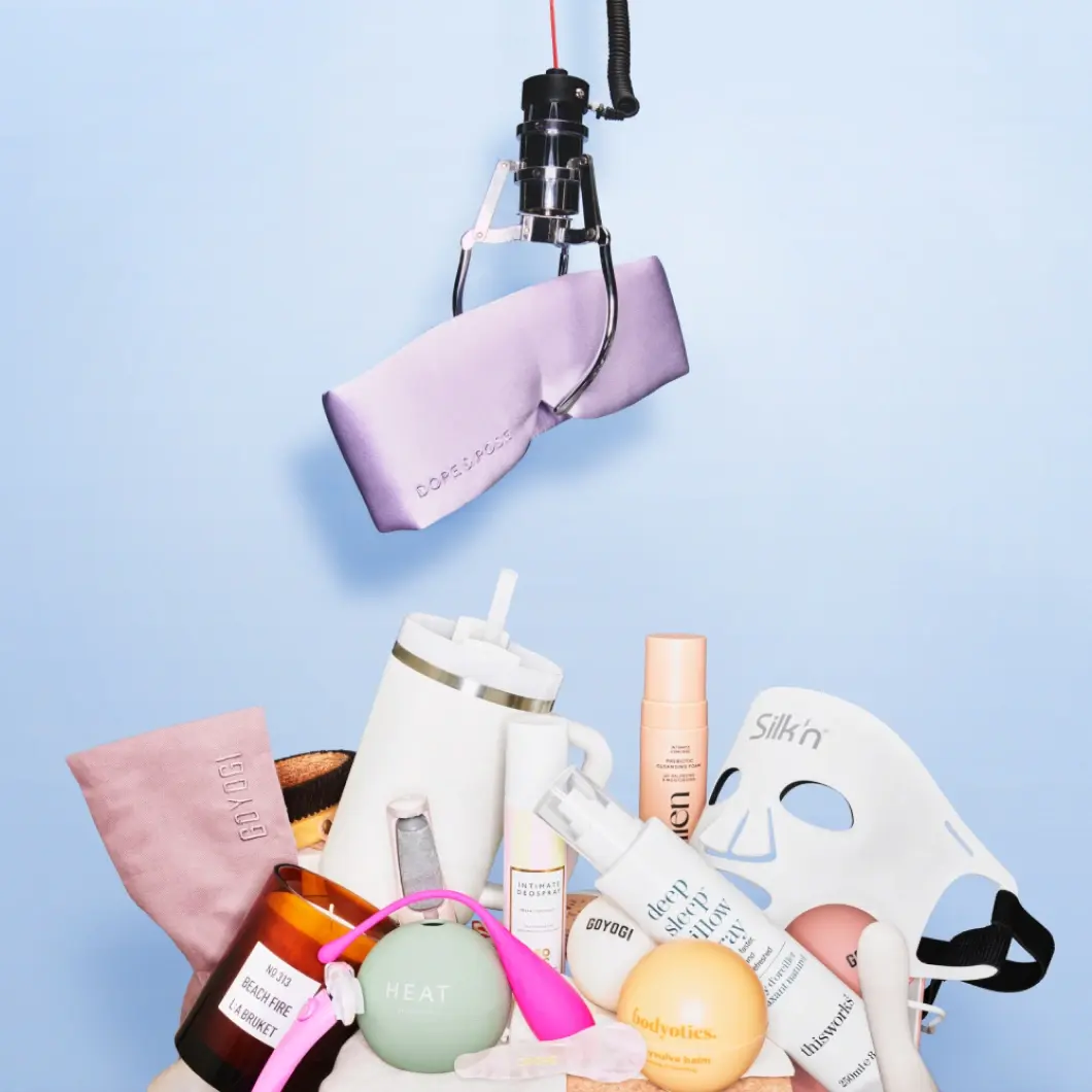 En kloarm holder et lilla silkelignende stoff over ulike wellnessprodukter mot lys blå bakgrunn. Produktene inkluderer Silkn LED-maske, drikkekopp, kroppspleie og wellness-tilbehør.