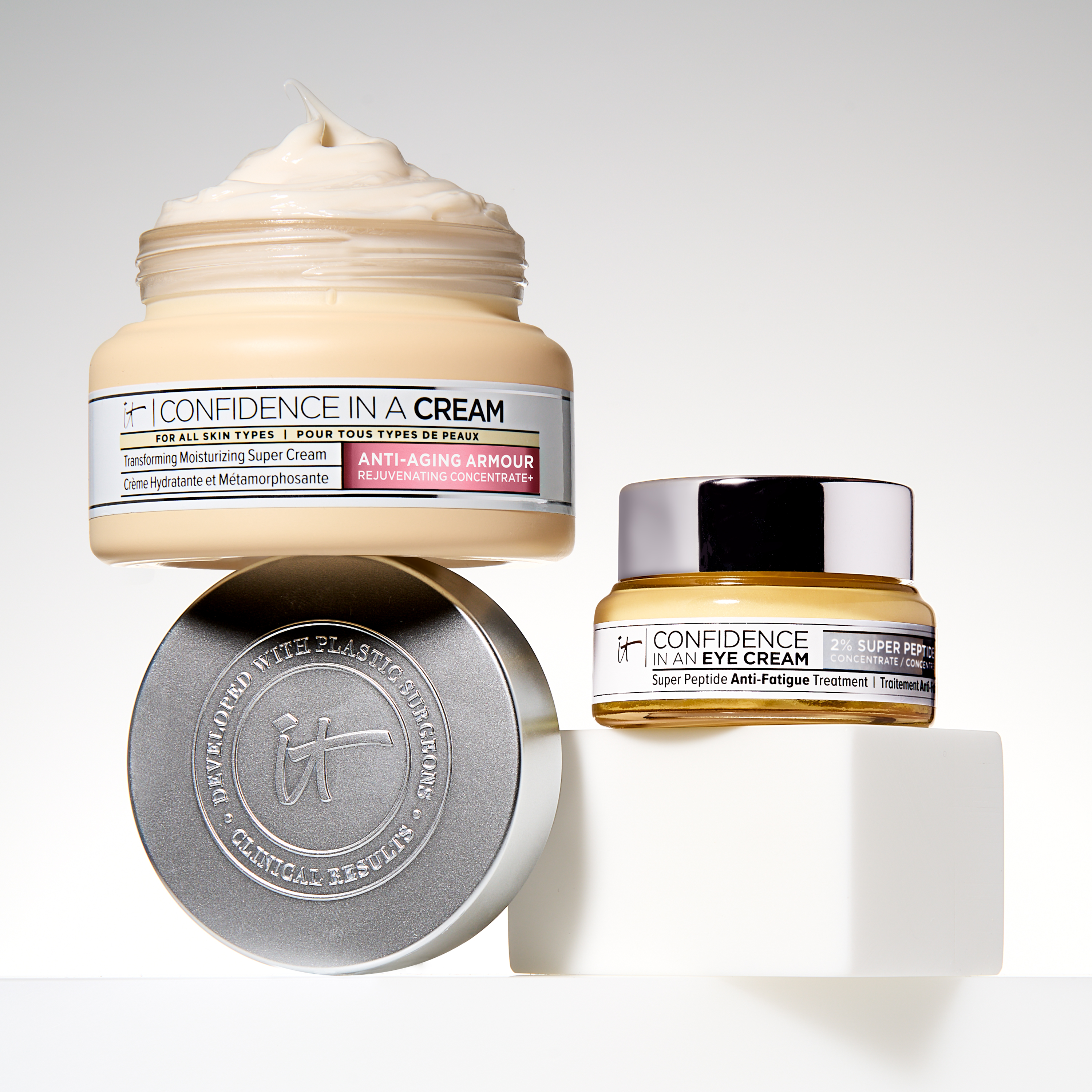 Kaksi ihonhoitotuotetta IT Cosmeticsilta: ”Confidence in a Cream” ja ”Confidence in an Eye Cream”. Tuotteet on sijoitettu valkoiselle pinnalle, jonka vieressä on hopeanvärinen kansi. Tausta on vaaleanharmaa.