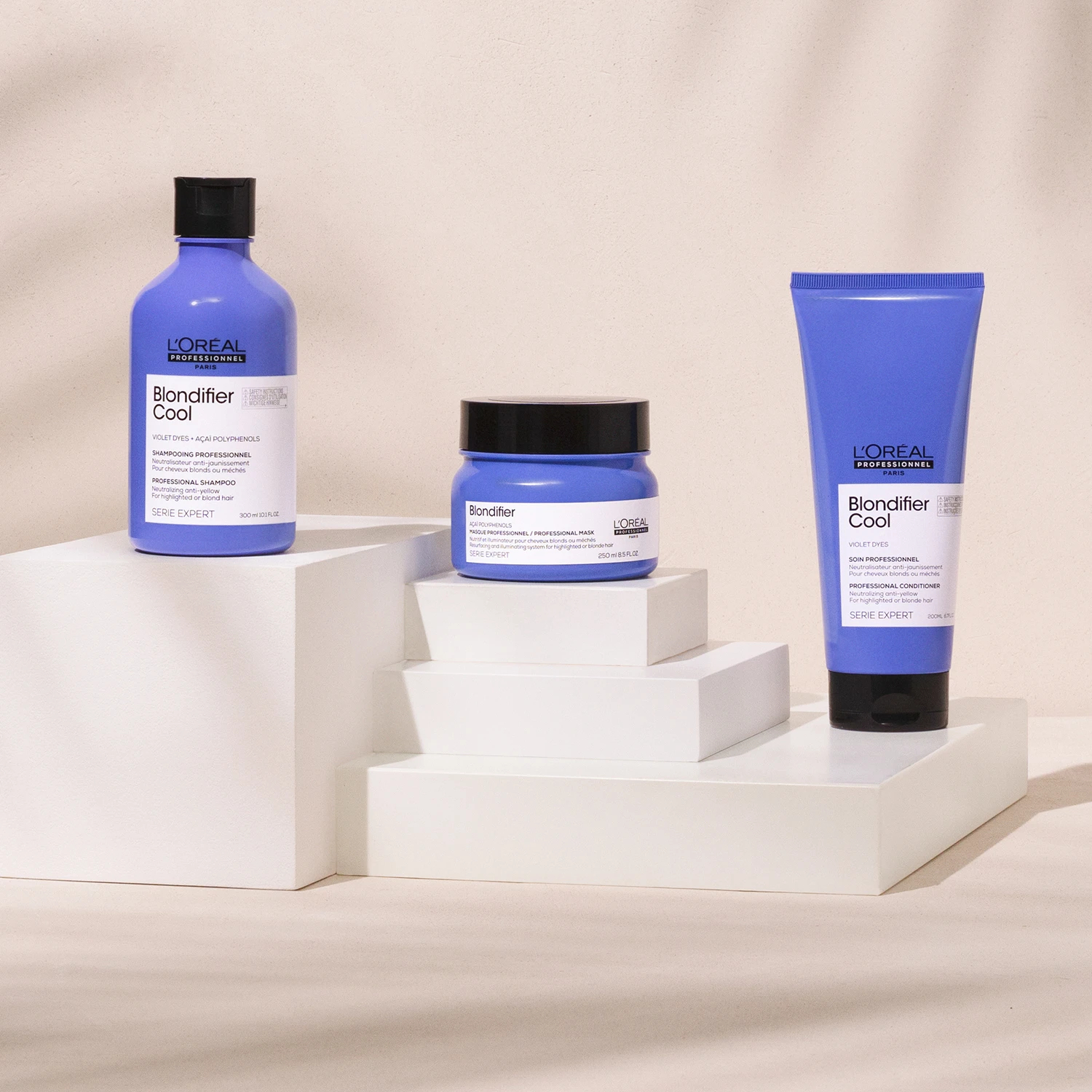 Tre hårvårdsprodukter från L'Oréal Professionnel Blondifier Cool-serien står uppradade på vita kuber mot en beige bakgrund. Alla produkter har blå förpackningar med vita etiketter och svarta lock.