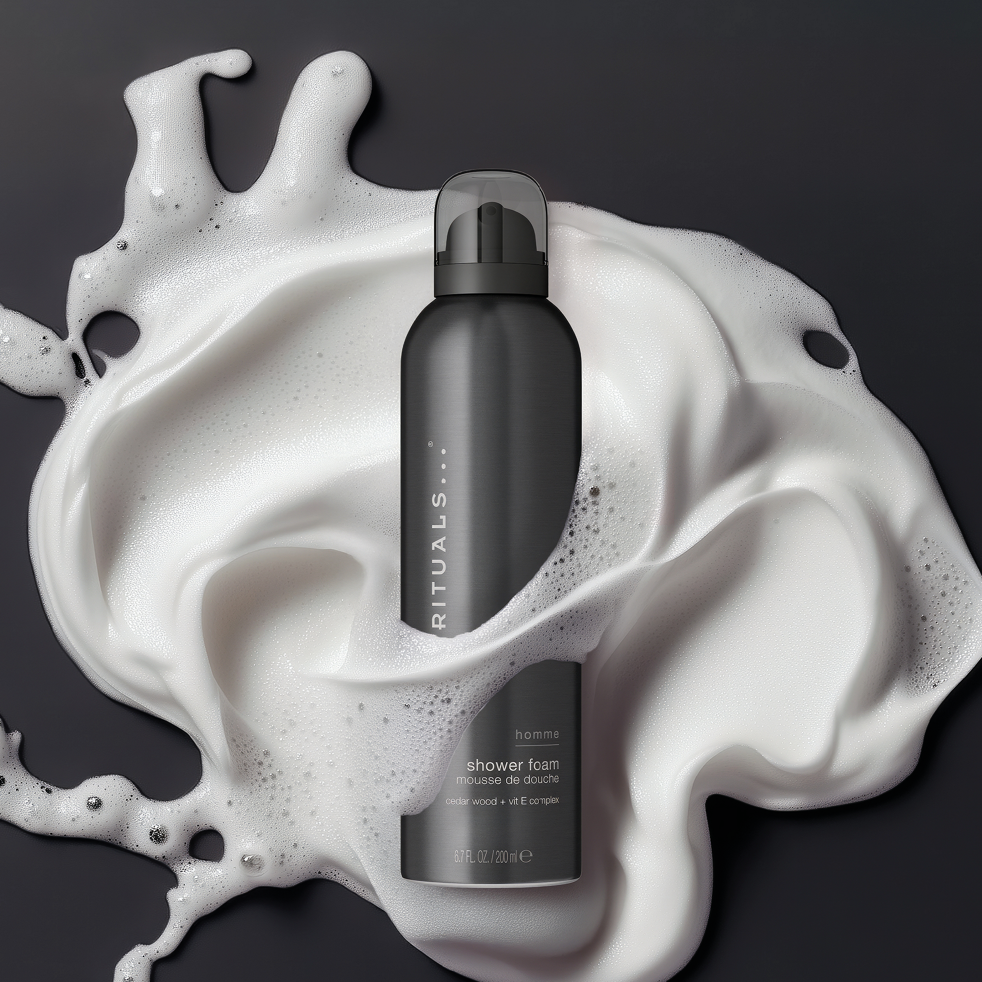 Musta Rituals Homme shower foam -pullo on valkoisen suihkuvaahdon keskellä tummalla taustalla. Pullossa näkyvät tekstit “RITUALS”, “homme”, “shower foam” ja “cedar wood + vit E complex”.