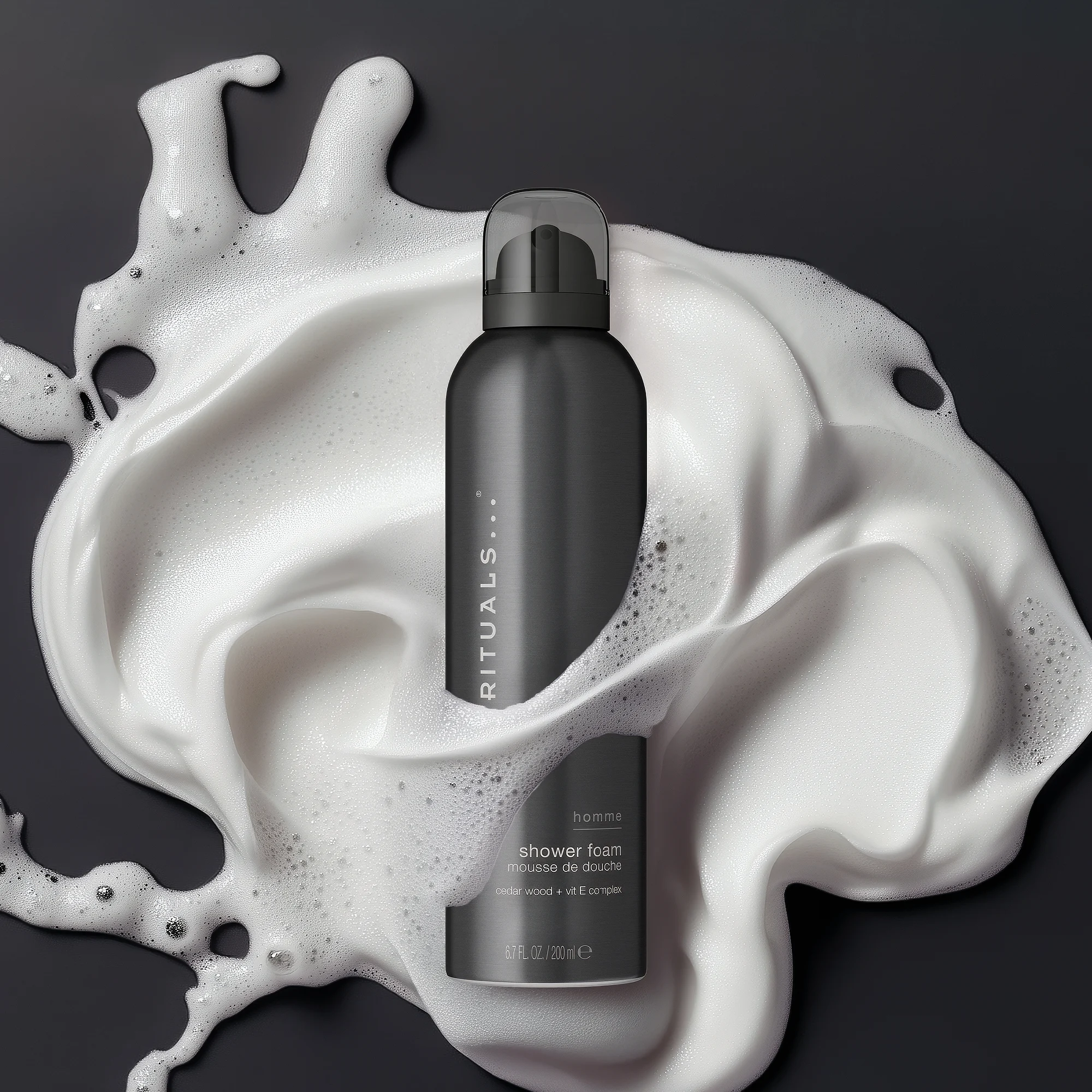 Svart flaska med Rituals Homme shower foam står i vit duschskum mot mörk bakgrund. På förpackningen syns texterna “RITUALS”, “homme”, “shower foam” och “cedar wood + vit E complex”.