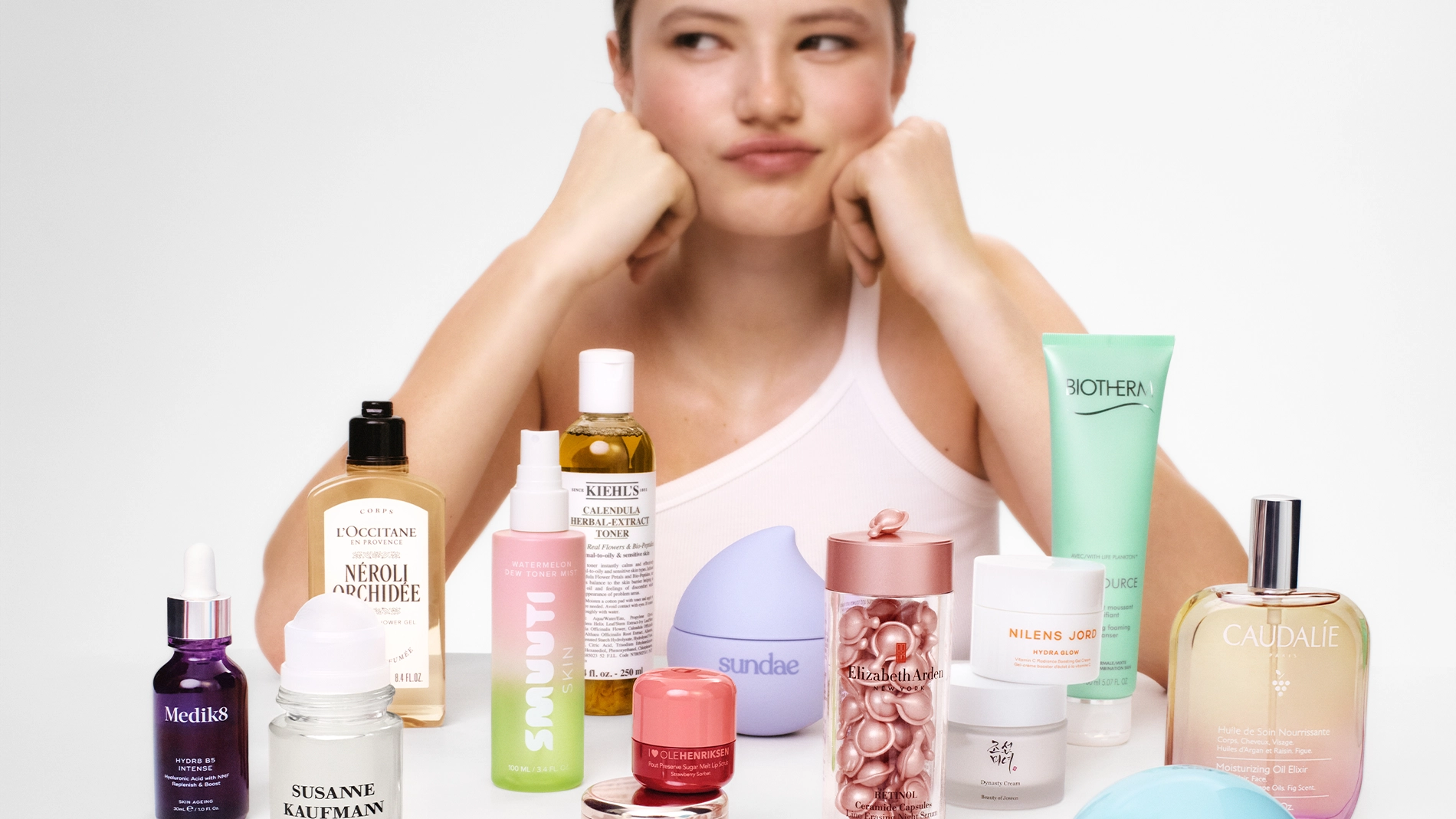 Person som lutar hakan i händerna bakom ett bord fyllt med hudvårds- och skönhetsprodukter, inklusive L’Occitane Néroli & Orchidée, Kiehl’s Calendula Herbal Extract Toner, Biotherm Aquasource, NUXE Huile Prodigieuse, Caudalie Eau des Vignes, Medik8 serum, Susanne Kaufmann Deodorant Roll-On och Elizabeth Arden kapselserum.