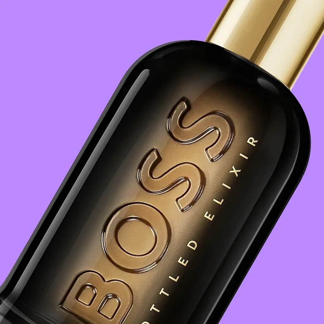Nærbilde av BOSS Bottled Elixir-parfyme i en svart og gullfarget flaske med gullfarget kork og synlig tekst «BOSS BOTTLED ELIXIR», mot lilla bakgrunn.