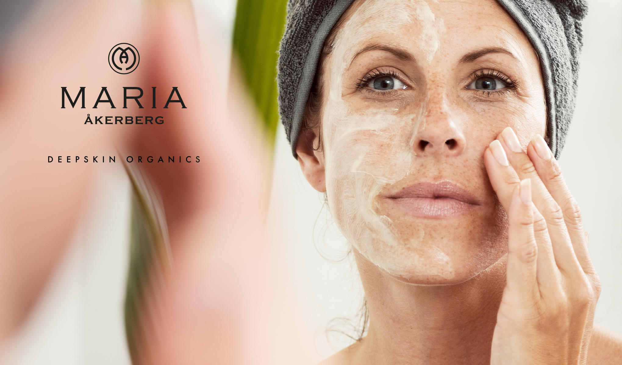 Henkilö, jolla on harmaa pyyhe päänsä ympärillä, levittää ihonhoitotuotetta kasvoilleen. Kuvan vasemmalla puolella on Maria Åkerbergin logo ja teksti ”Deepskin Organics”. Tausta on vaalea ja epäselvä.