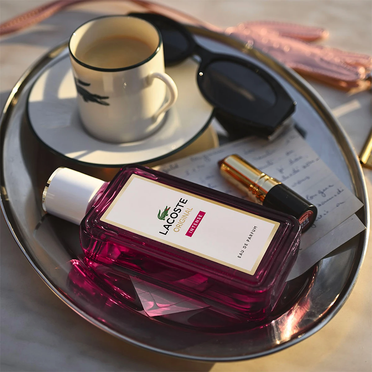 Purppura Lacoste Original Intense Eau de Parfum -pullo, kahvikuppi, aurinkolasit ja huulipuna hopeatarjottimella.