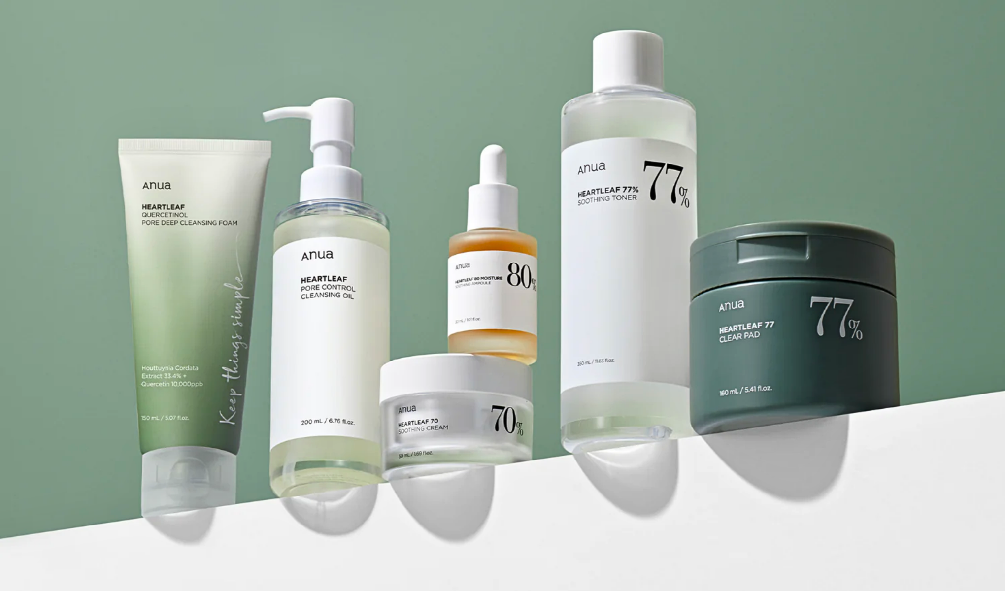 En serie hudpleieprodukter fra merket Anua. Produktene er plassert på en hvit flate med grønn bakgrunn og har en minimalistisk innpakning med hvite og grønne farger og enkel tekst.