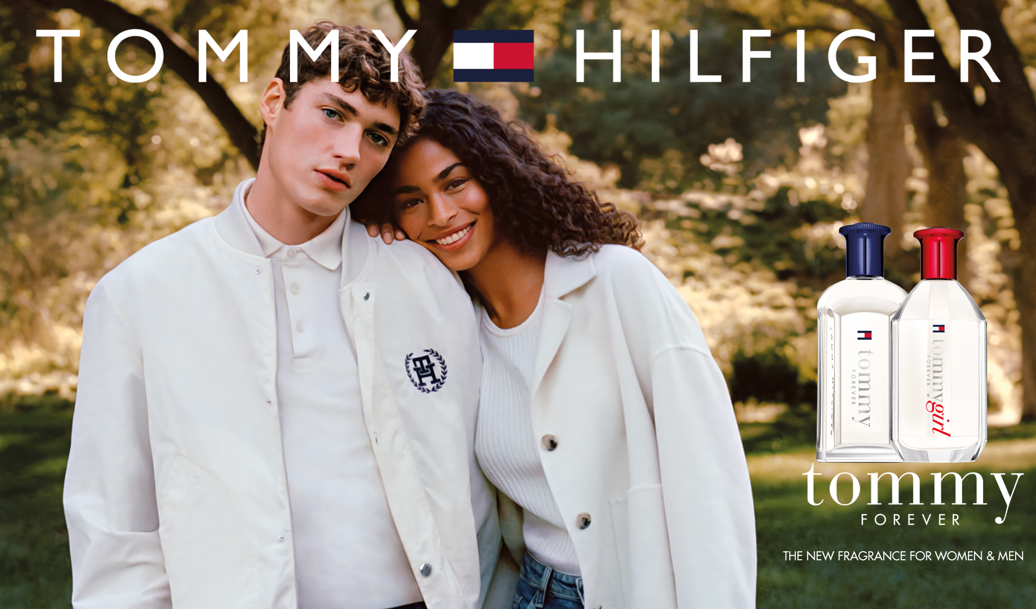 Kaksi valkoisiin vaatteisiin pukeutunutta ihmistä seisoo lähellä toisiaan vihreässä puistossa. Ylhäällä on teksti ”Tommy Hilfiger” ja tuotemerkin logo. Oikealla alhaalla on kaksi hajuvesipulloa, joissa lukee ”Tommy Forever” ja ”Tommy Girl Forever” sekä sanat ”The new fragrance for women & men”.