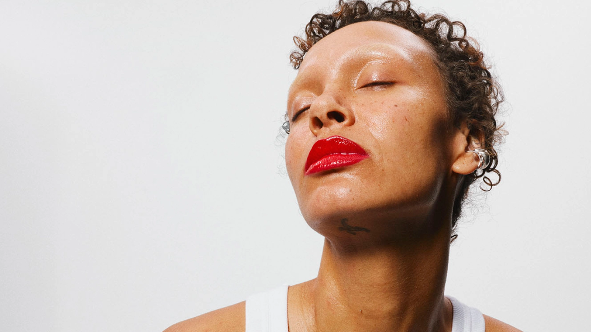 Puoliprofiilinen muotokuva henkilöstä vaaleaa taustaa vasten. Henkilöllä on lyhyet kiharat hiukset, suljetut silmät ja punainen huulipuna. Hartiat ja ylävartalon osa näkyvät, ja henkilöllä on valkoinen hihaton paita.