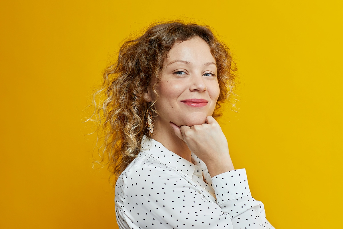 Person med lockigt hår poserar framför gul bakgrund med naturlig makeup och glansiga rosa läppar. Vit prickig blus och hängande örhängen skapar en modern beauty- och hårstylinglook.