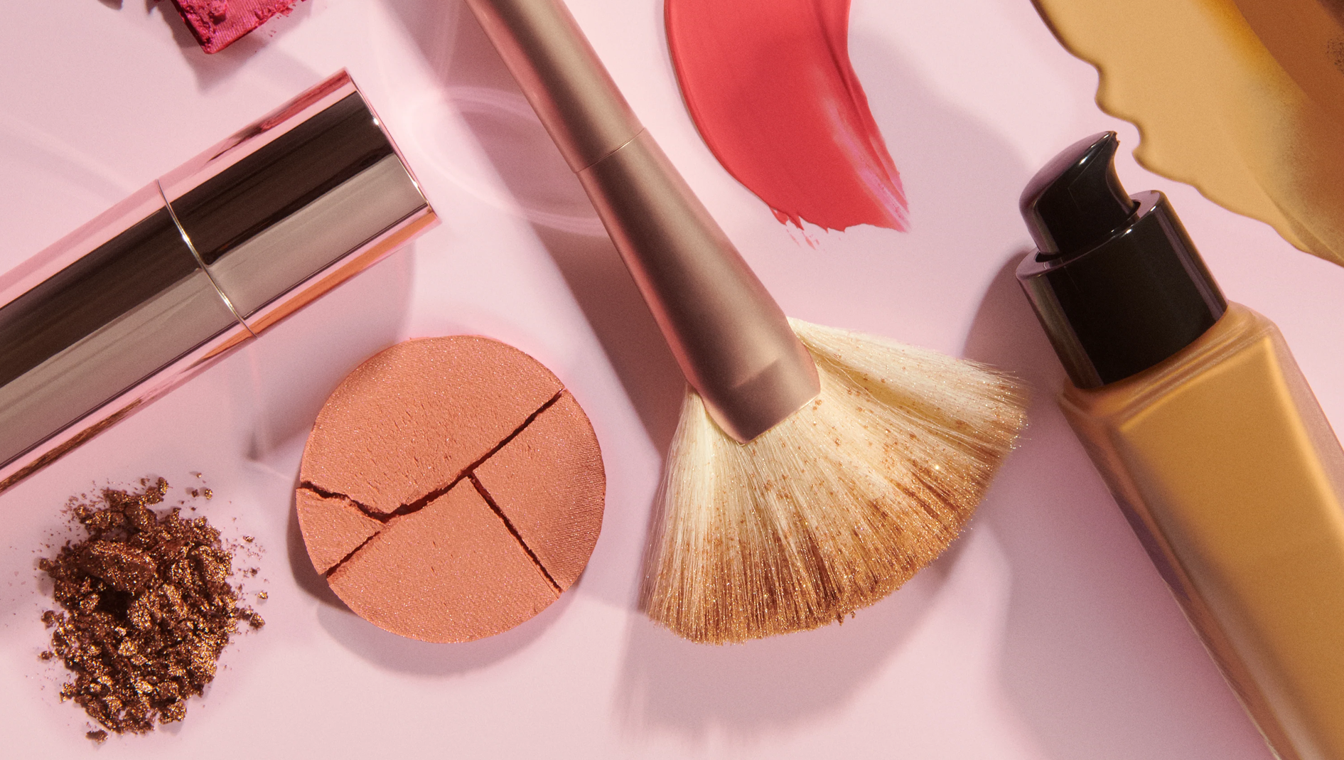Makeupprodukter på rosa bakgrund med foundation i pumpflaska, sminkborste, läpprodukt, krossat puder och färgprover i rosa och guld.