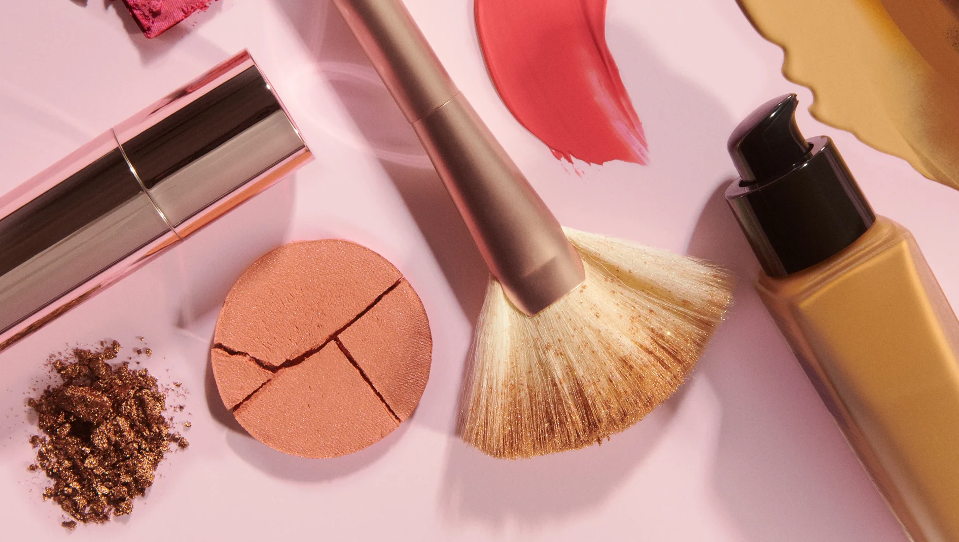 Makeupprodukter på rosa bakgrund med foundation i pumpflaska, sminkborste, läpprodukt, krossat puder och färgprover i rosa och guld.