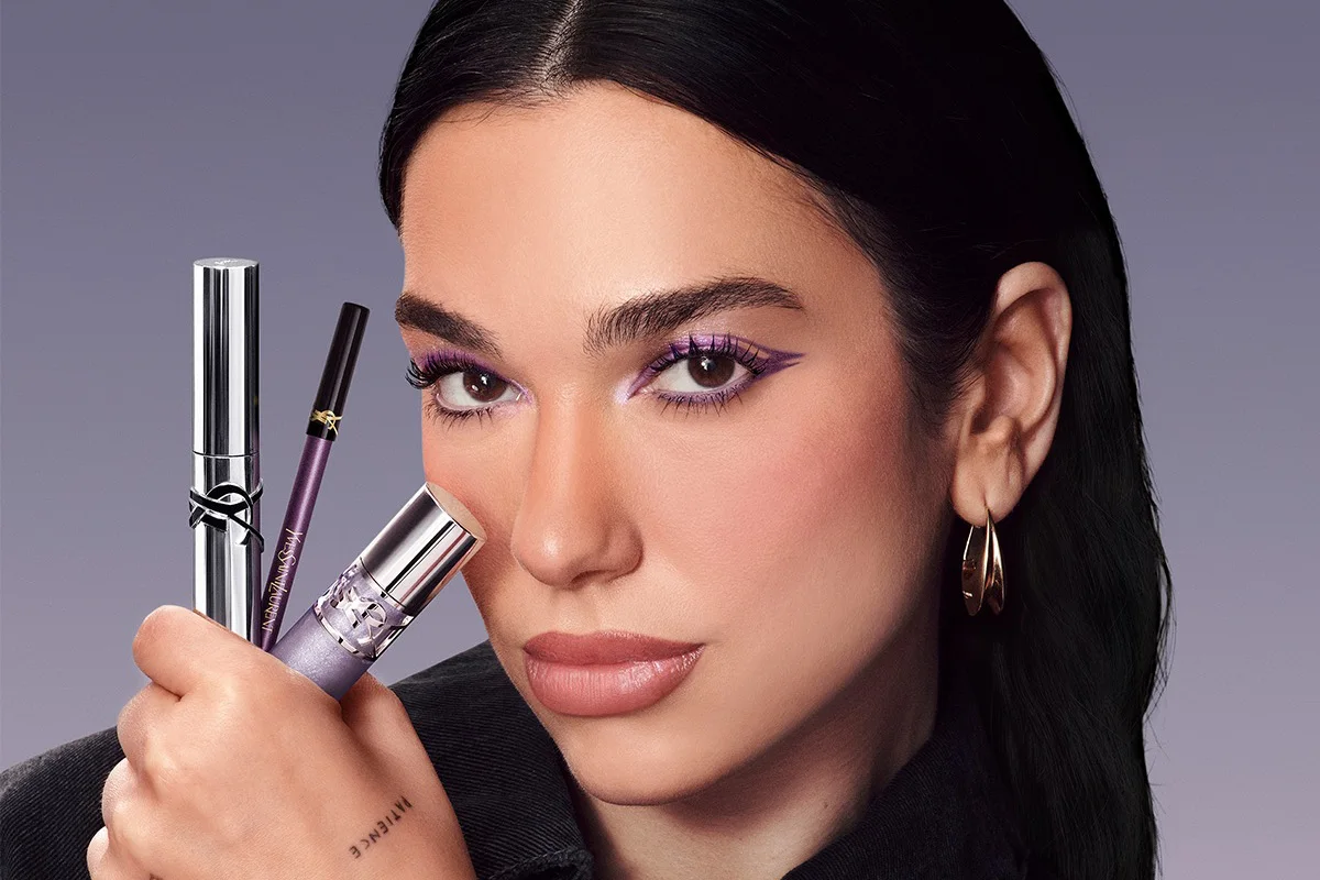 En person bär lila ögonmakeup och håller tre Yves Saint Laurent Beauty-sminkprodukter framför ansiktet: en mascara i silverfärgad hylsa, en lila eyeliner och en concealer med lila förpackning. Produkterna visar YSL-logotypen och texten "Yves Saint Laurent".