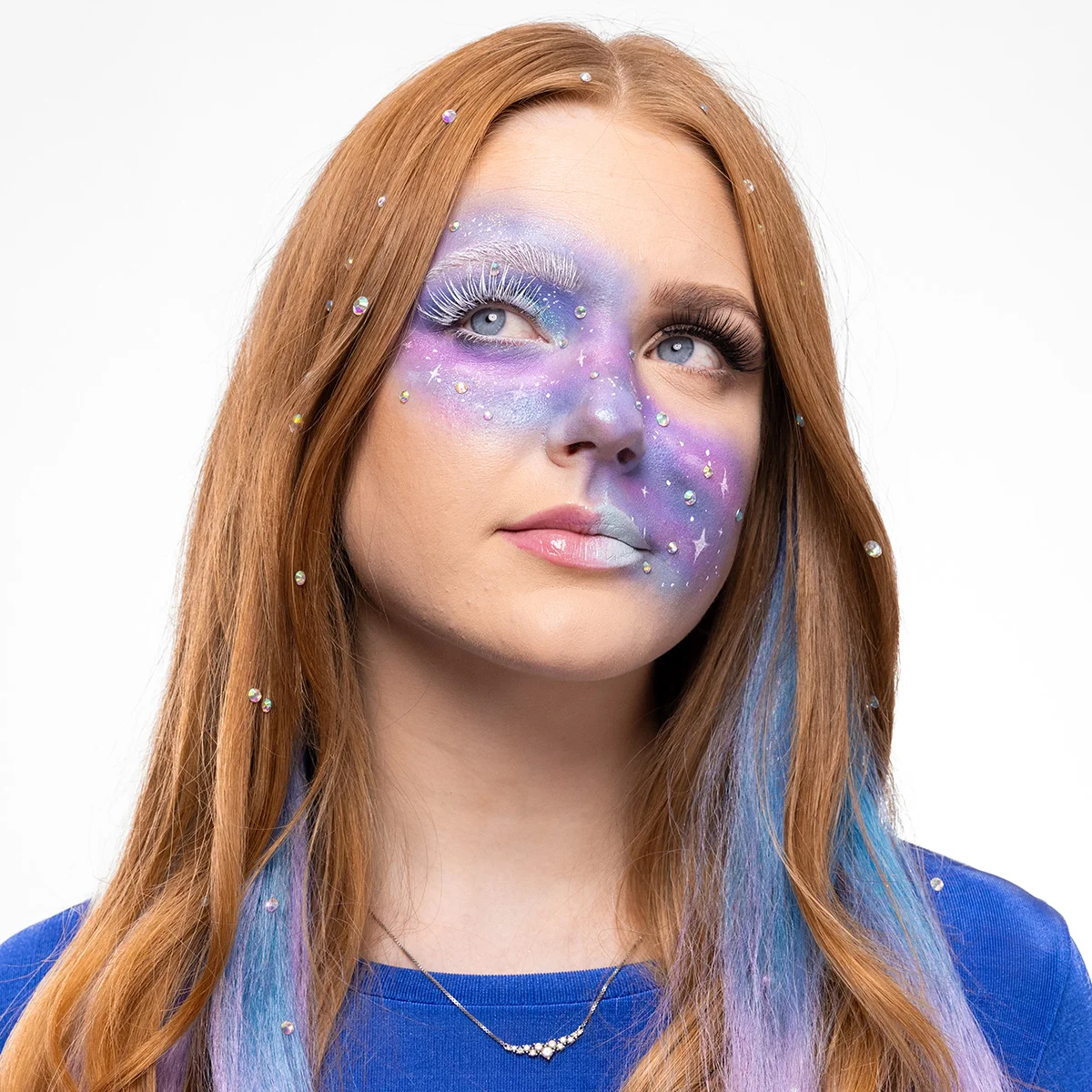 En person med galaxinspirerad makeup i lila, blå och silvertoner med vita fransar, små glittrande stenar och stjärnliknande detaljer över ansiktet och i håret.