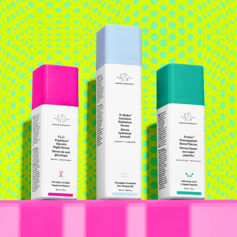 Tre Drunk Elephant-serum i firkantede flasker med fargede lokk (rosa, blått og turkis) mot en grønn og gul mønstret bakgrunn.