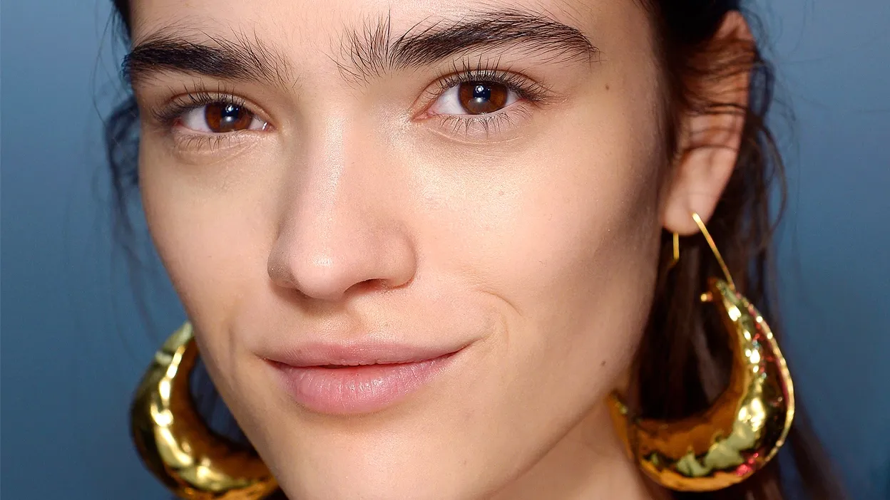 En person med glatt hud, mørke øyne og dristige gulløreringer som smiler diskret.