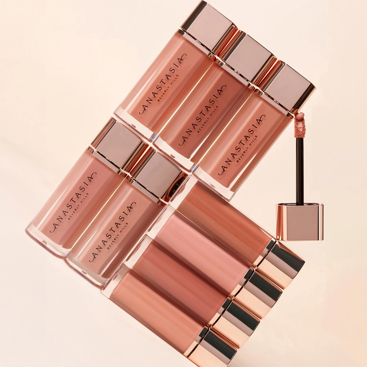Bilde av åtte lipglosser fra Anastasia Beverly Hills i ulike nyanser av rosa og nude, arrangert i et geometrisk mønster. En av lipglossapplikatorene er forlenget og synlig.