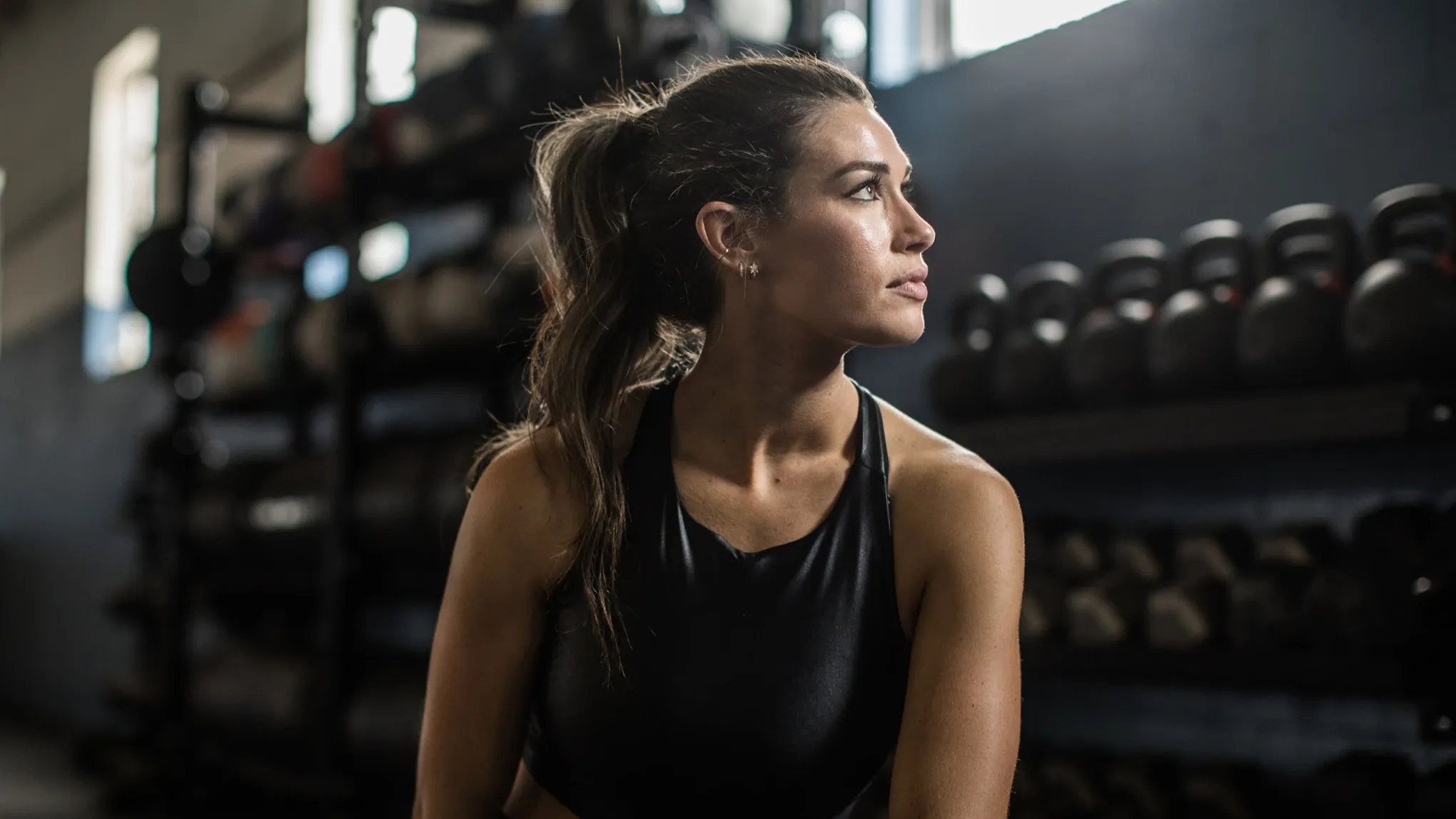 Kvinne i treningsklær sitter på treningssenter og ser til siden, med kettlebells i bakgrunnen.