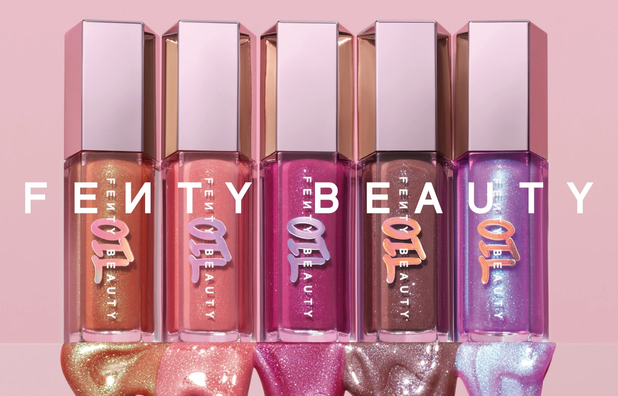 Fenty Beautyn uusin kokoelma erilaisia kimaltelevia huulikiiltoja on tyylikkäästi aseteltu vaaleanpunaista taustaa vasten. Jokaisella kiillolla on ainutlaatuinen sävy ja hohtava viimeistely, täydellinen antamaan huulille säihkyvän ilmeen.