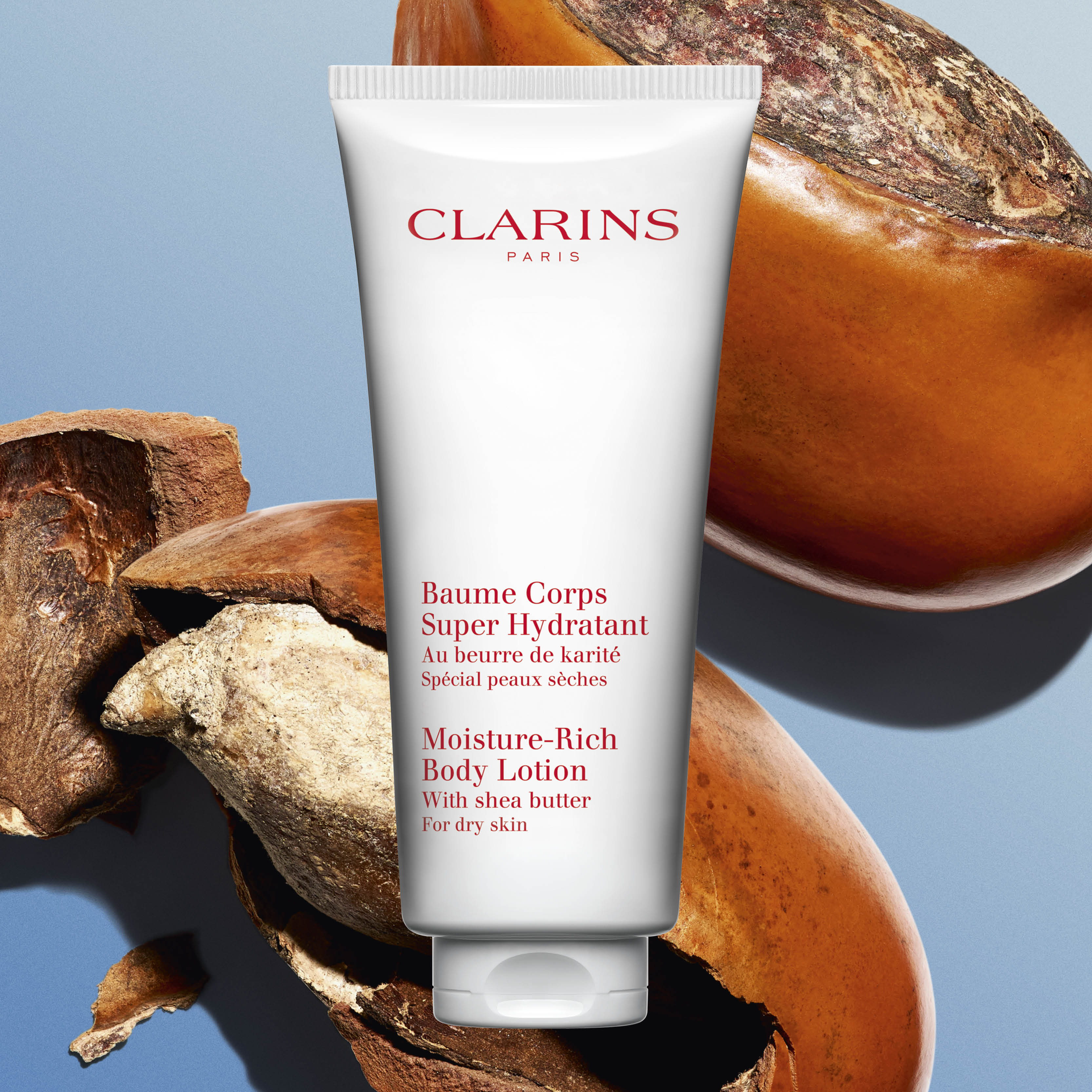 Clarinsin tuote ”Baume Corps Super Hydratant” , taustalla sheapähkinöitä. Tuotteen teksti on sekä ranskaksi että englanniksi, ja se kuvaa tuotetta kosteuttavaksi vartalovoiteeksi kuivalle iholle.