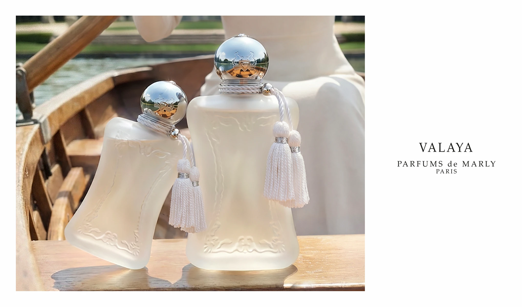 Kvinne i hvitt holder Parfums de Marly Valaya-parfyme i en hage ved siden av en stor hvit blomst.