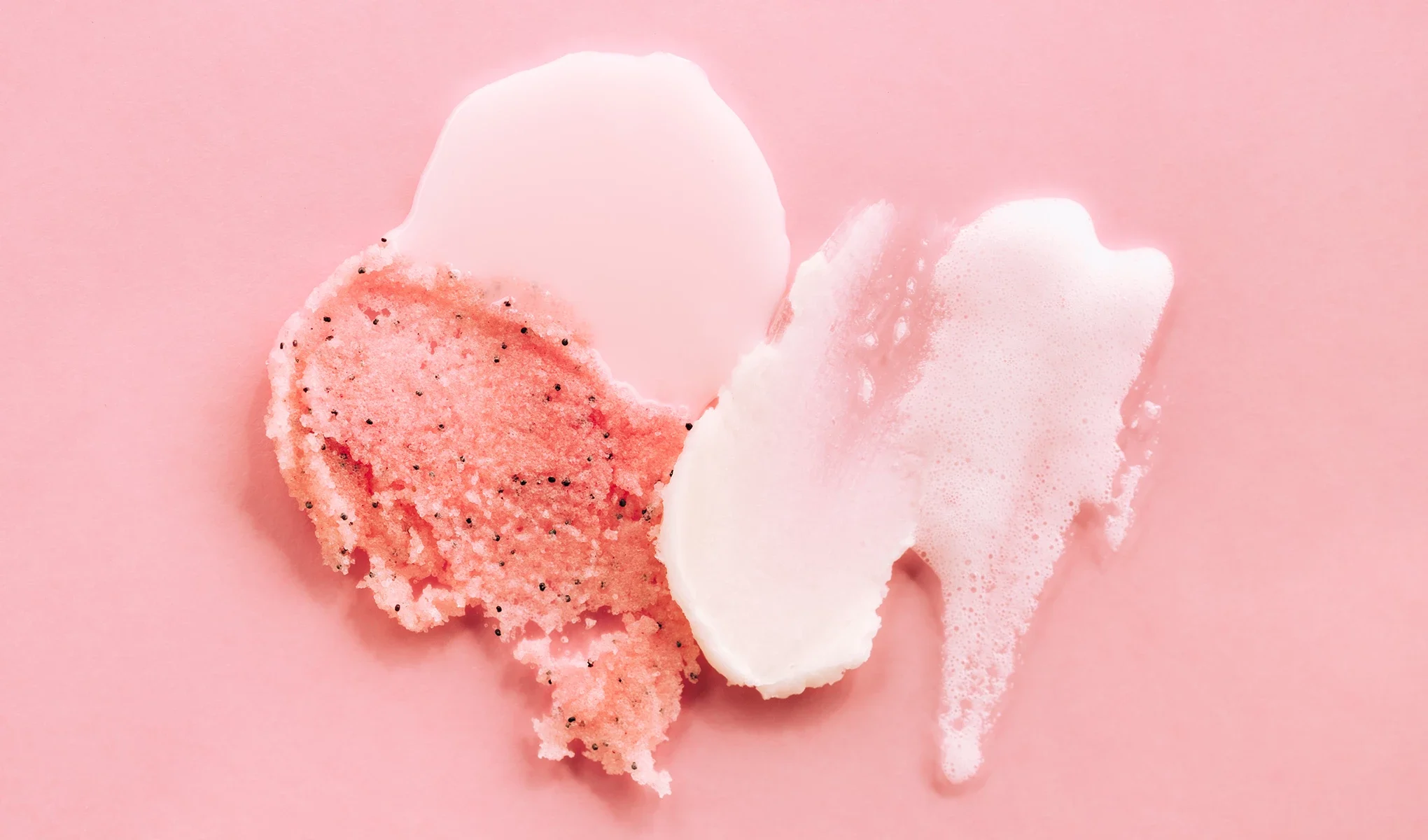 Ulike teksturer av ansiktspeeling, inkludert gelé, skrubb og skum, i rosatoner på en rosa bakgrunn