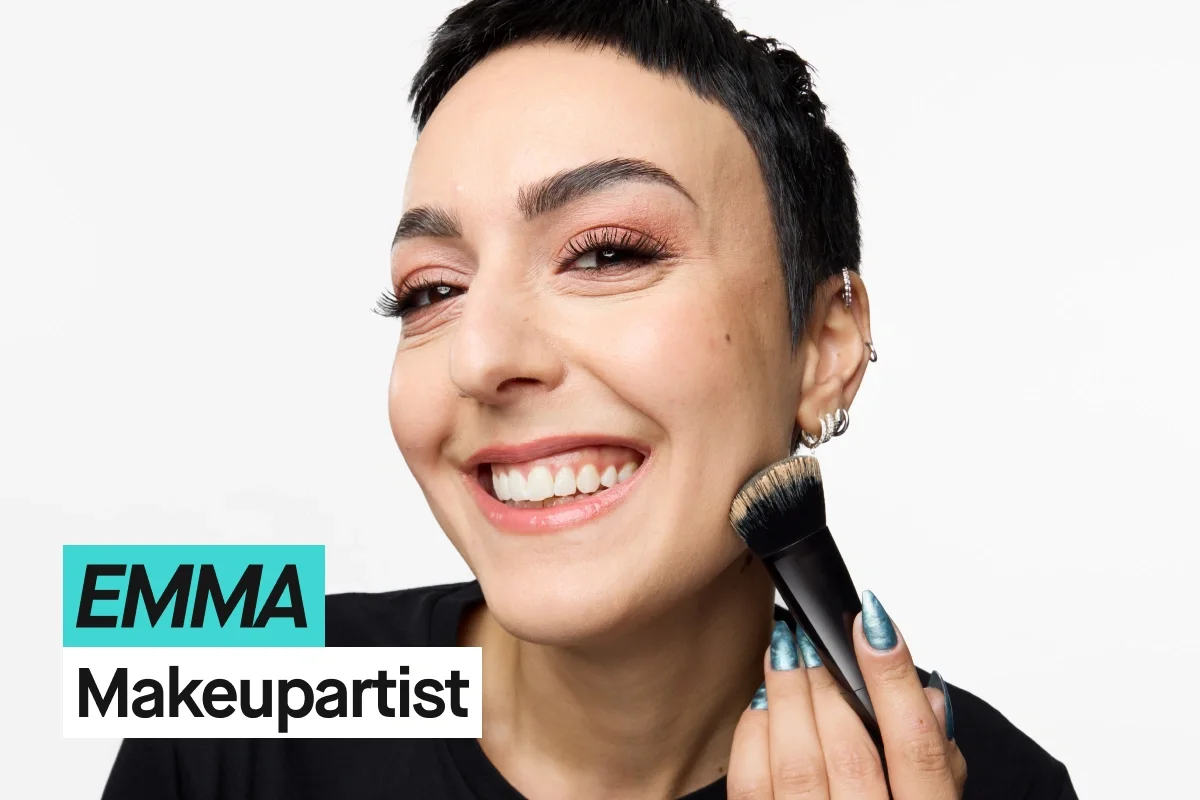 Person med kort svart hår påfører makeup med sminkekost mot kinnet og har på seg en svart KICKS-t-skjorte mot lys grå bakgrunn. Turkis og hvit tekst viser “EMMA” og “Makeupartist”.