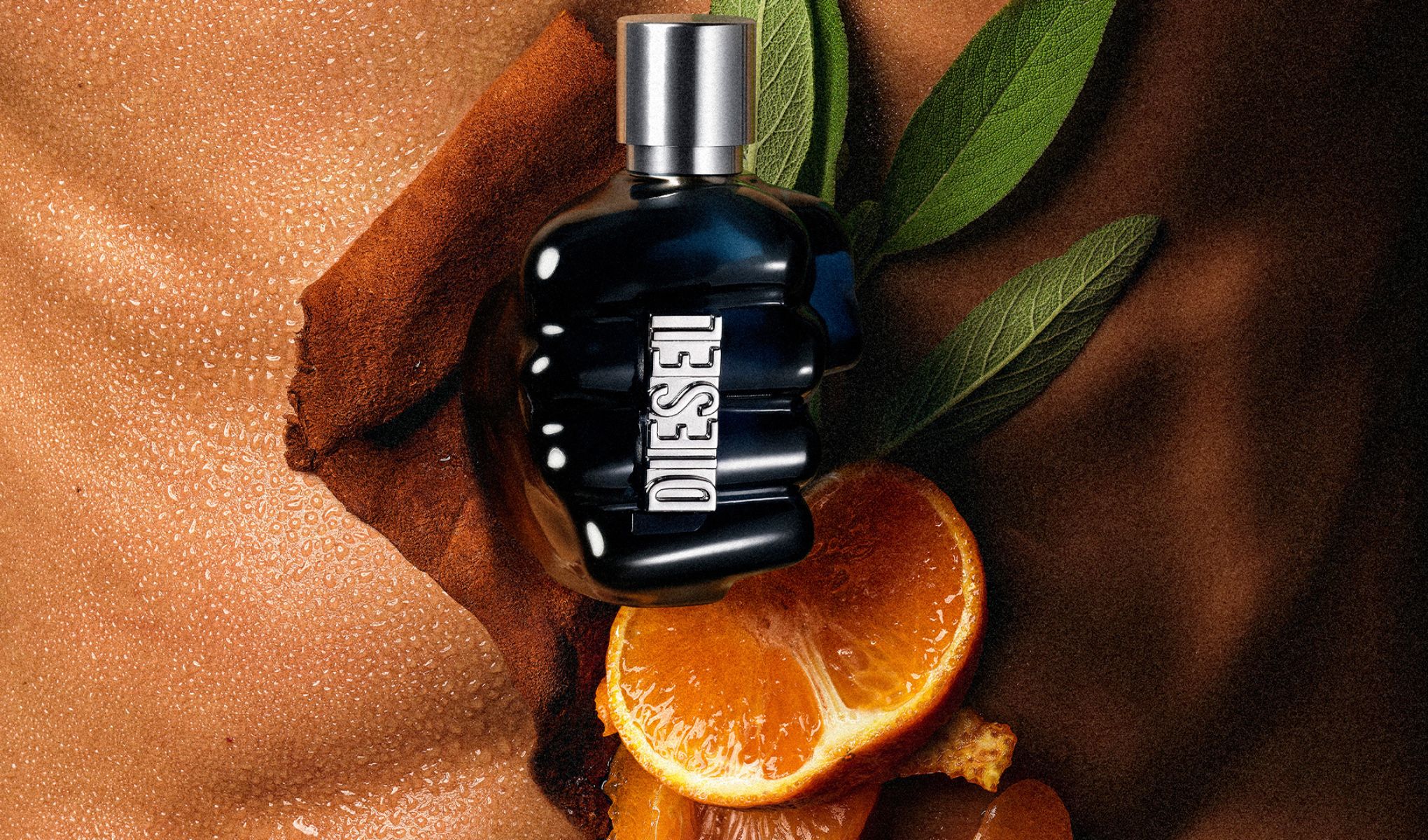 Svart DIESEL parfymeflaske med sølvlokk ligger på en overflate med kanelpulver, appelsinskiver og grønne blader. Glassflasken med mørk væske er omgitt av ingredienser som fremhever duftens karakter.