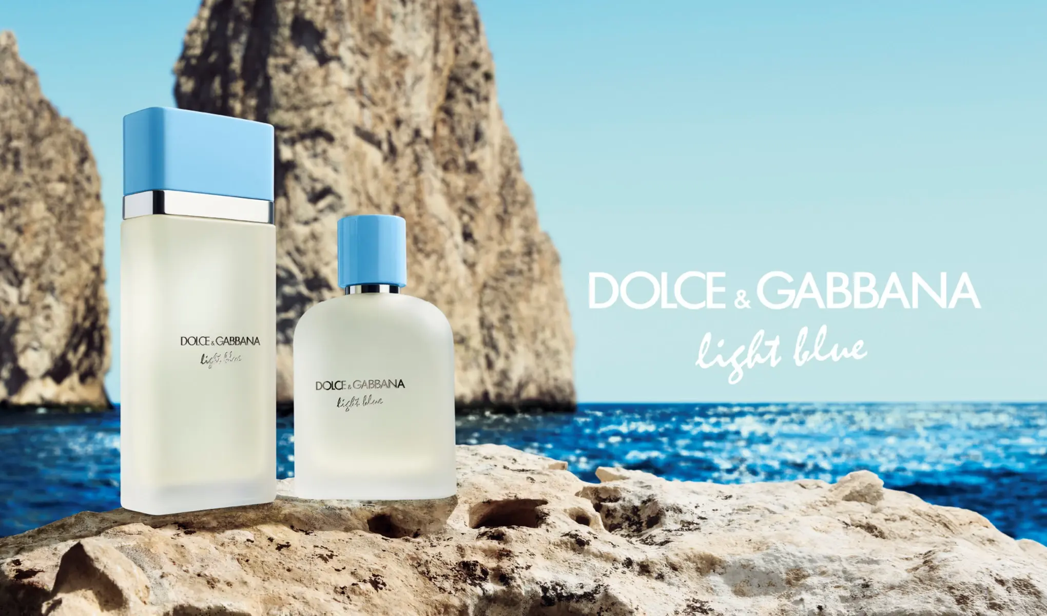 Två parfymflaskor från Dolce & Gabbana Light Blue står på en sten framför ett blått hav med klippor i bakgrunden. Flaskorna har ljusblå korkar och en minimalistisk design.