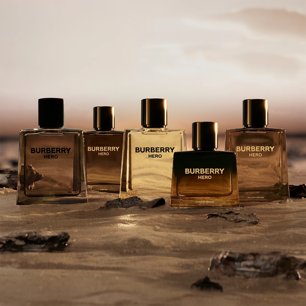 Fem Burberry Hero-doftflaskor placerade på sand vid vattenkanten i solnedgång.