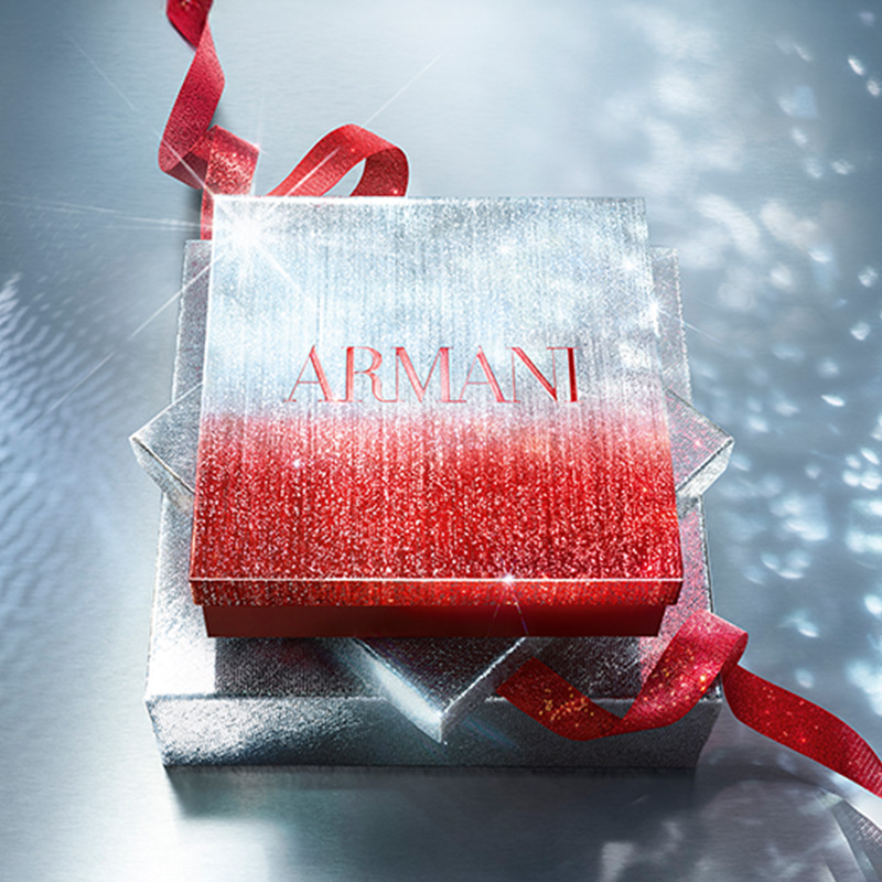 På en sølvfarget overflate ligger to gaveesker med rød og sølv glitteroverflate. Eskene er dekorert med rødt bånd og har teksten "Armani".