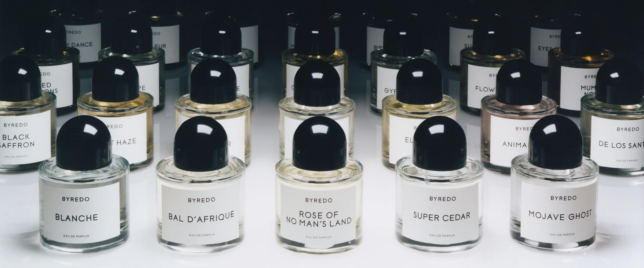 Flera parfymflaskor från Byredo, arrangerade på en vit yta. Varje flaska har en svart kork och en vit etikett med parfymens namn, såsom "Black Saffron", "Blanche", "Bal D'afrique", "Rose Of No Man's Land", "Super Cedar" och "Mojave Ghost".