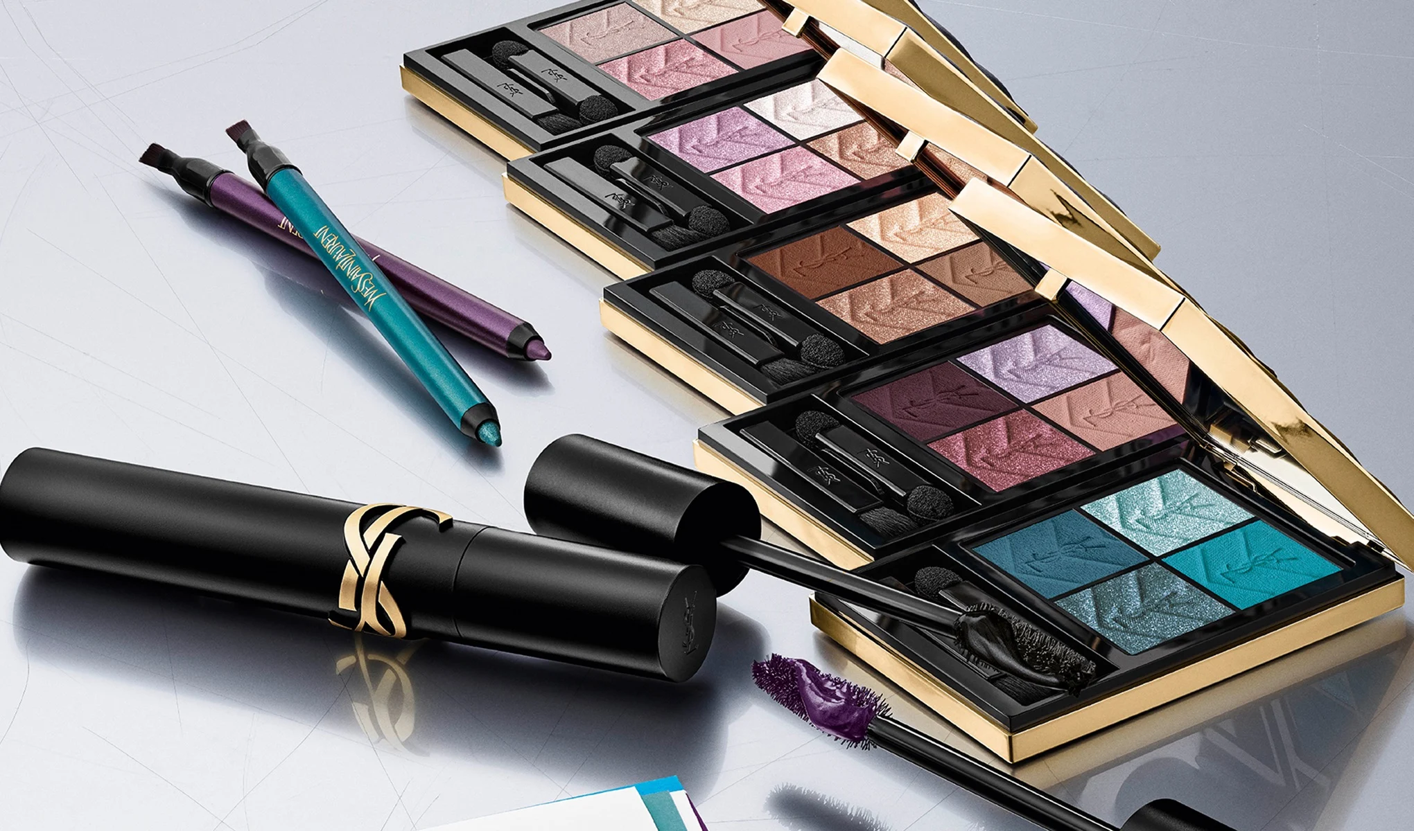  Yves Saint Laurent-sminkeprodukter arrangert på lys bakgrunn. Fire åpne øyenskyggepaletter med applikatorer vises i blå, lilla, rosa og brune nyanser sammen med to eyelinerpenner i turkis og lilla med YSL-logo samt svart mascara med gullfarget YSL-detalj.