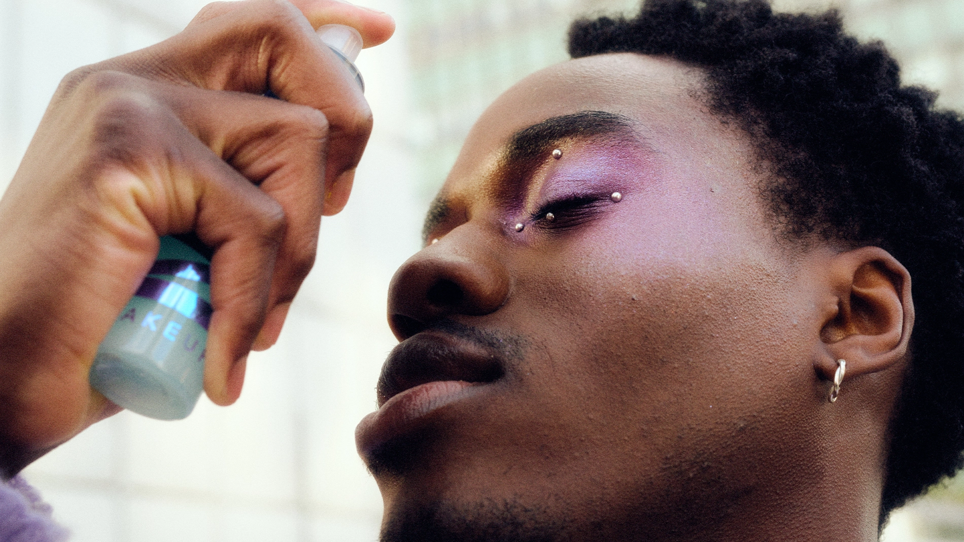 Person med kort hår og fargerik øyenskygge i lilla og gult sprayer settingspray mot ansiktet. Nærbilde i profil med lukkede øyne, to piercinger i øyenbrynet og øredobb i øret. Flaske med ansiktsspray holdes tett mot huden mot lys bakgrunn.