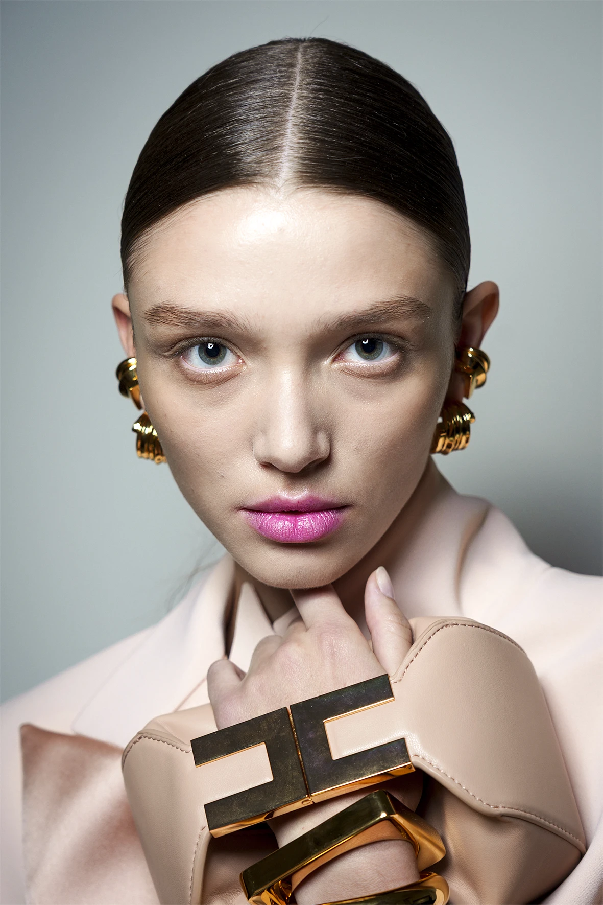 Närbild på en person med slät hud och mittbena, som bär rosa läppstift och stora guldfärgade örhängen samt armband. Personen håller handen mot hakan och bär en beige jacka, med fokus på makeup som läppstift och naturlig hudfinish.