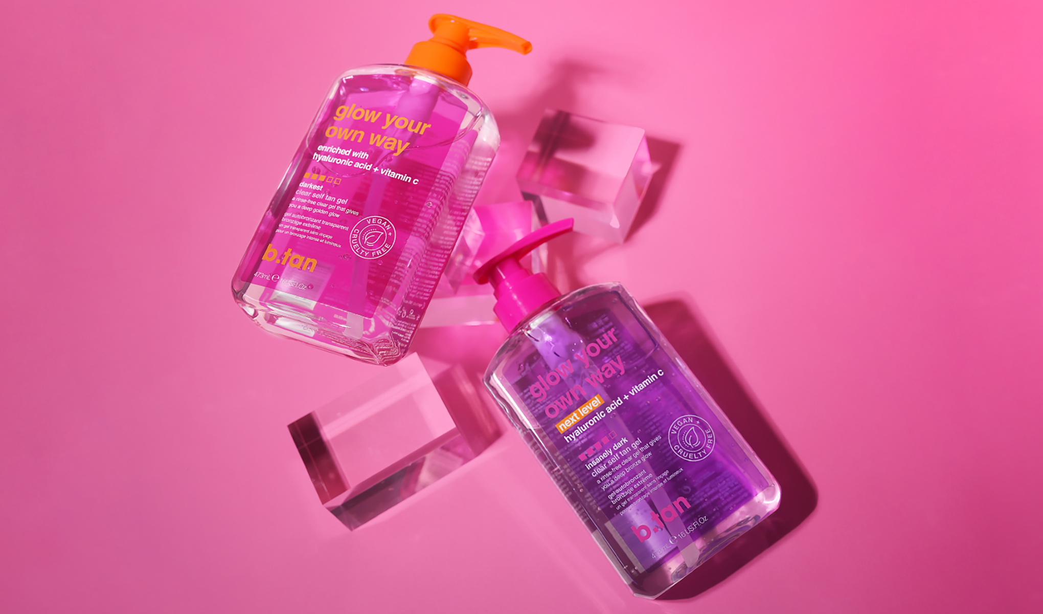 To flasker med b.tan solgelé er plassert på en rosa bakgrunn. Den ene flasken har en oransje pumpe, og den andre har en rosa pumpe. Flaskene er gjennomsiktige og inneholder gel med hyaluronsyre og C-vitamin. Teksten på flaskene lyder «glow your own way».