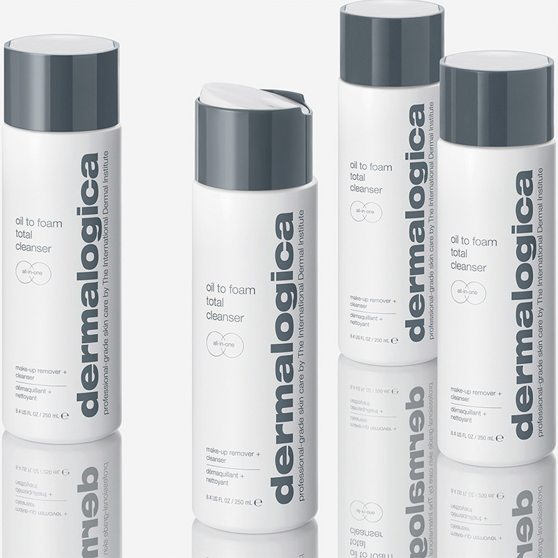 Fyra flaskor av produkten "Oil to Foam Total Cleanser" från märket Dermalogica. Flaskorna är vita med grå lock och har texten "dermalogica" tryckt vertikalt längs sidan. Flaskorna står på en reflekterande yta som skapar en spegeleffekt. Bakgrunden är ljus och neutral.