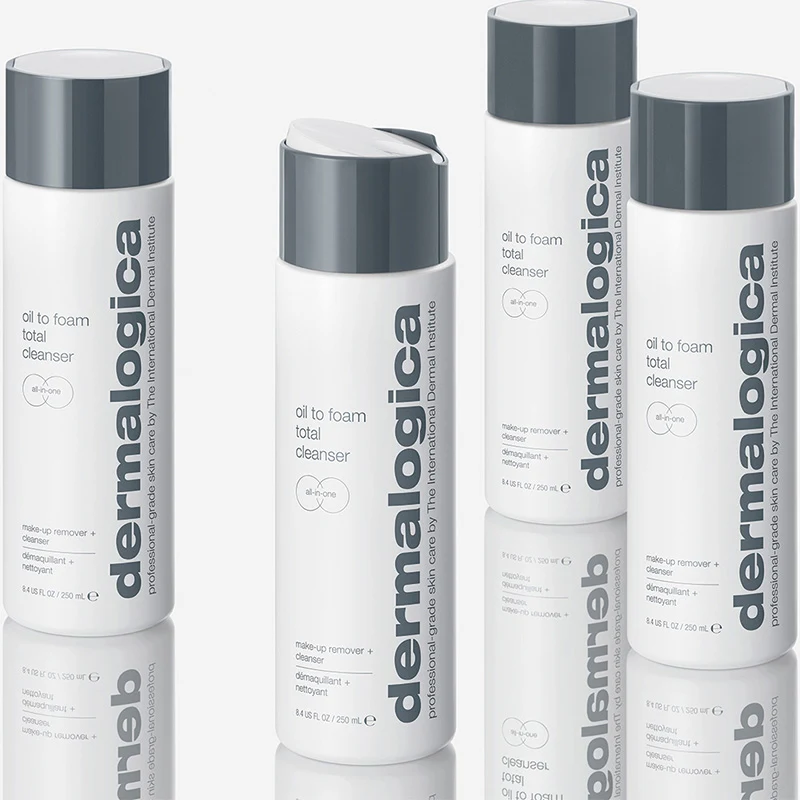 Fyra flaskor av produkten "Oil to Foam Total Cleanser" från märket Dermalogica. Flaskorna är vita med grå lock och har texten "dermalogica" tryckt vertikalt längs sidan. Flaskorna står på en reflekterande yta som skapar en spegeleffekt. Bakgrunden är ljus och neutral.