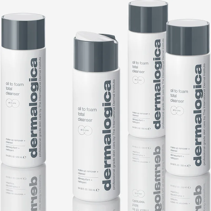 Fyra flaskor av produkten "Oil to Foam Total Cleanser" från märket Dermalogica. Flaskorna är vita med grå lock och har texten "dermalogica" tryckt vertikalt längs sidan. Flaskorna står på en reflekterande yta som skapar en spegeleffekt. Bakgrunden är ljus och neutral.