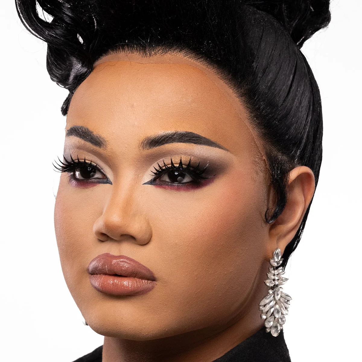 Närbild av en person med mörkt hår uppsatt i en elegant frisyr. Personen bär ett stort, glittrande örhänge och är klädd i svart.