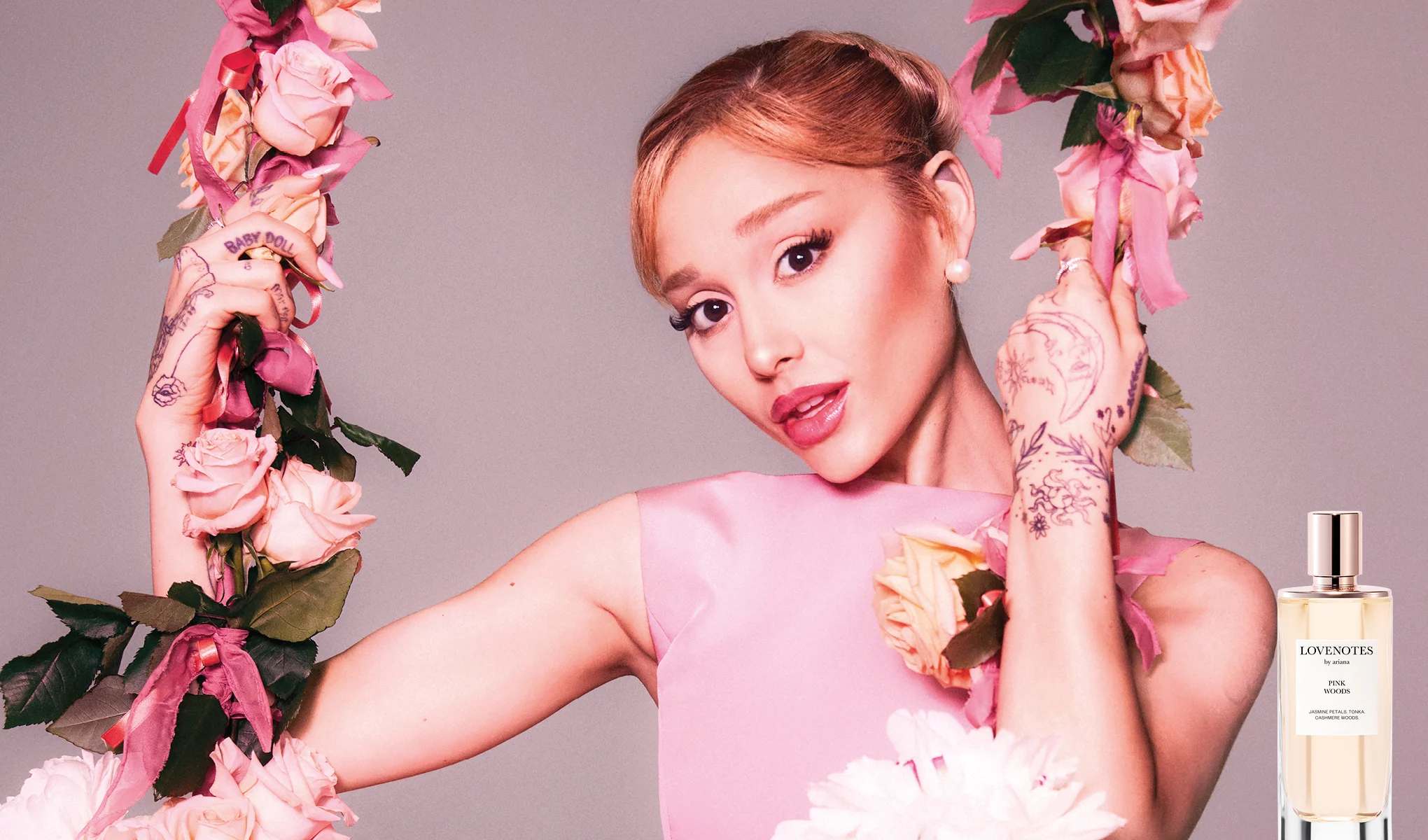 Ariana Grande keinussa vaaleanpunaisten kukkien keskellä ja Lovenotes Pink Woods -hajuvesipullo.