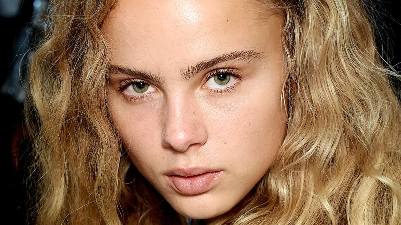 Person med vågigt blont hår och gröna ögon som tittar rakt in i kameran. Huden är klar och ser naturlig ut, med minimal eller ingen makeup.