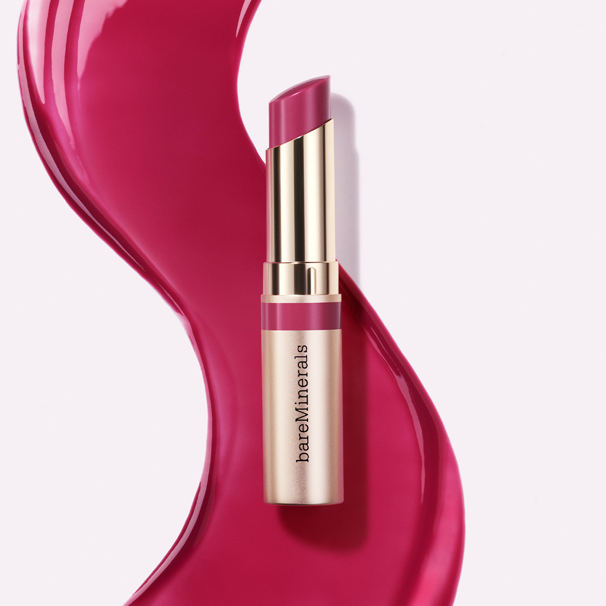 Leppestiften er rosa og har en gullfarget hylse med teksten «bareMinerals» på siden. Bakgrunnen er hvit med en stor, bølgende rosa fargeflekk som går diagonalt over bildet.