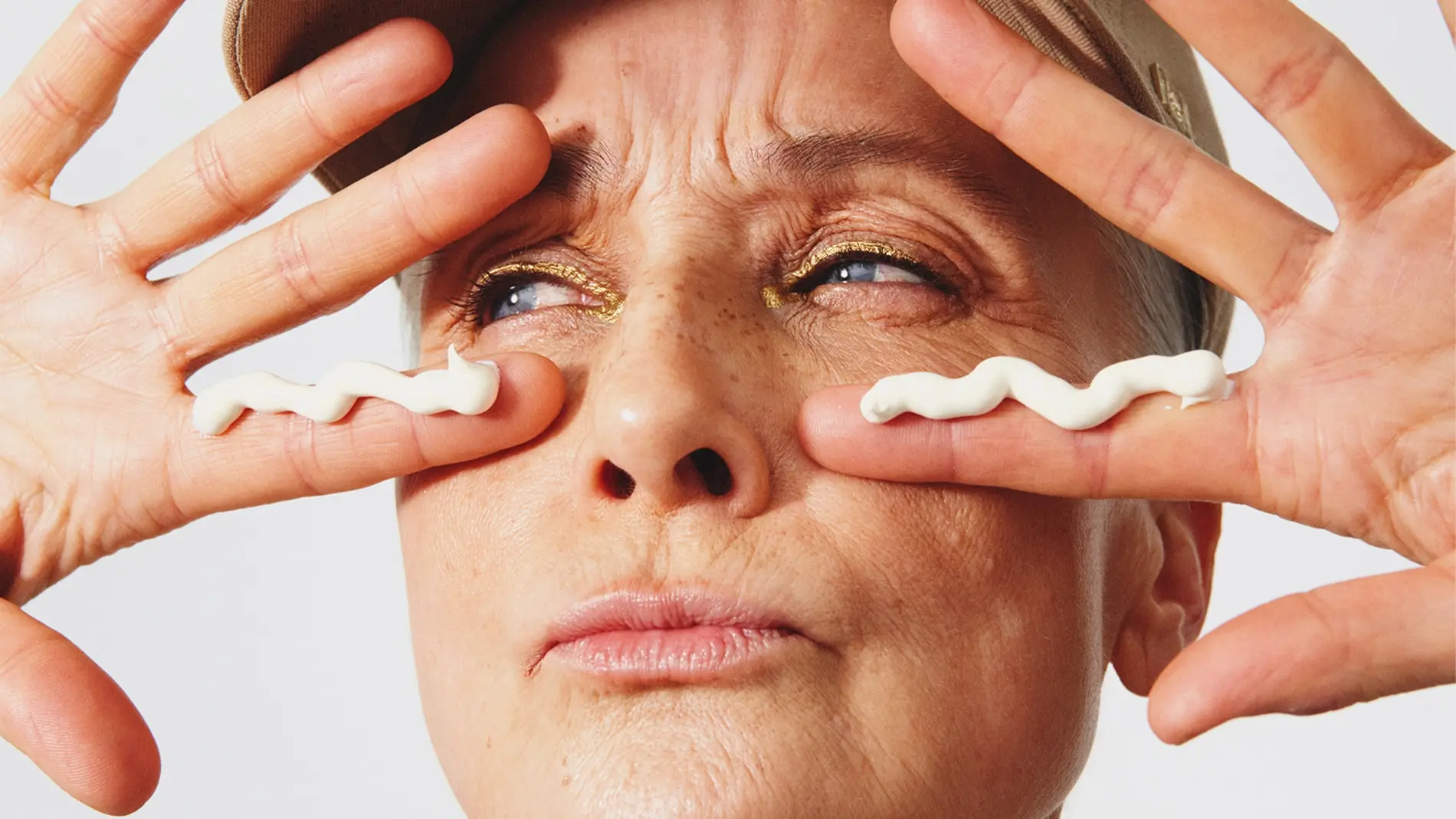 Henkilöllä on päässään beige lippalakki, ja hän pitää käsiään kasvojensa edessä, samalla kun silmien alle on levitetty kaksi valkoista juovaa ihonhoitovoidetta. Kullanhohtoinen luomiväri erottuu selvästi, ja lähikuva korostaa ihonhoidon levitystä vaaleaa taustaa vasten.