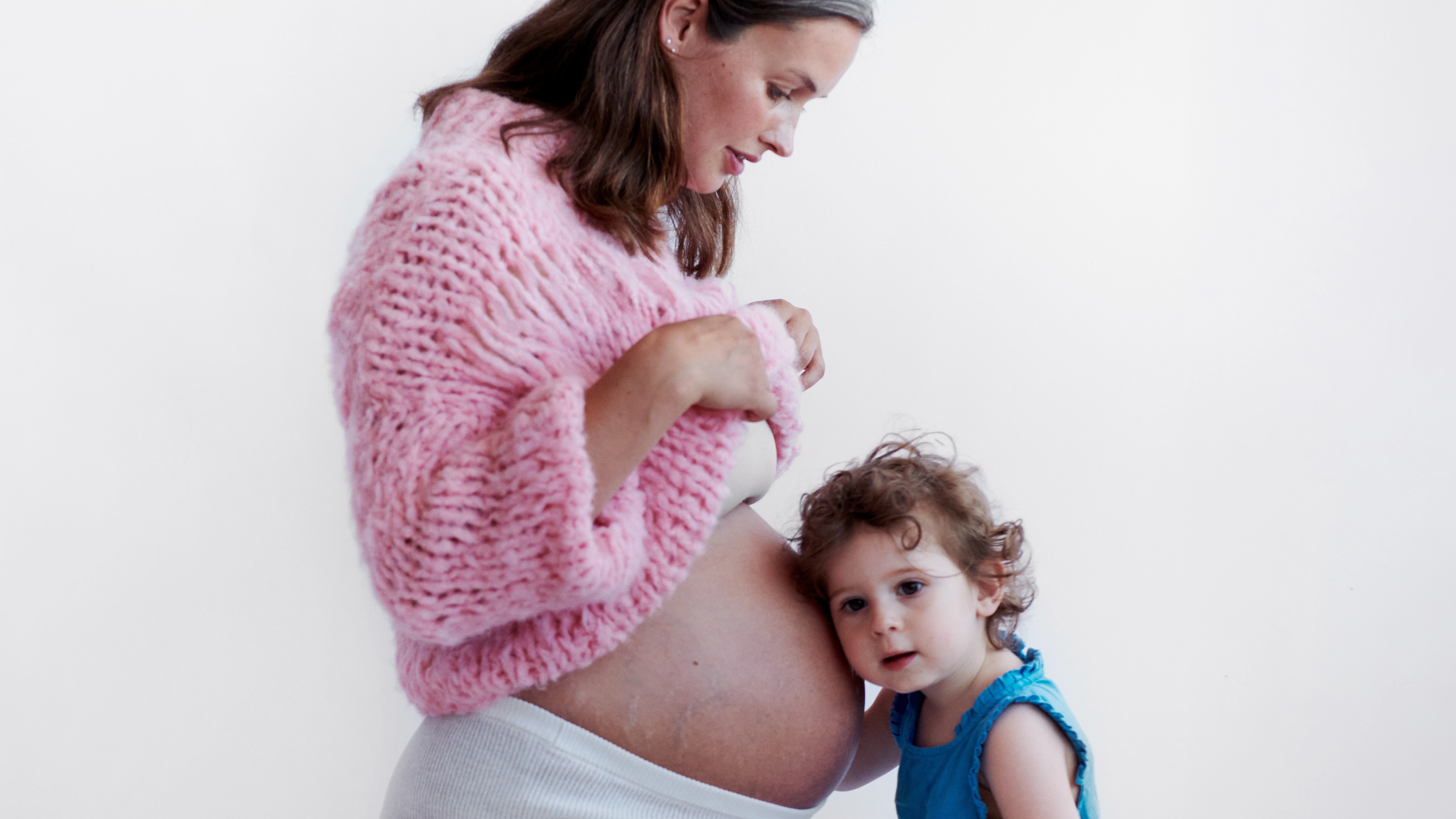 En gravid person knæler iført lyserød sweater, mens et barn i blå kjole lytter til den bare mave.