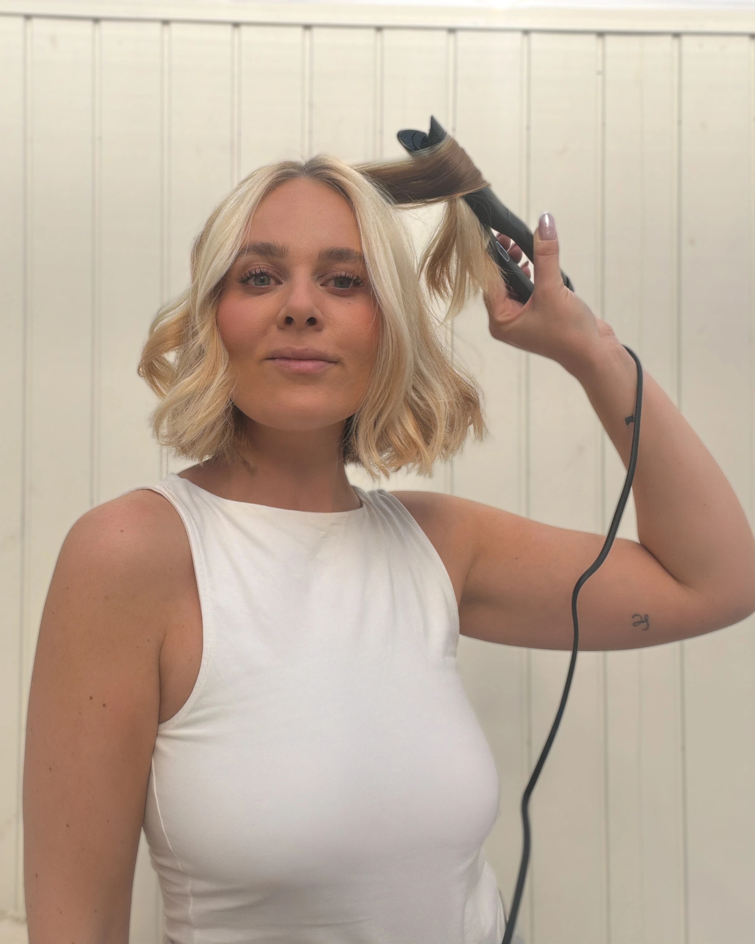 Kvinde styler sit blonde hår med et krøllejern og kigger ind i kameraet.