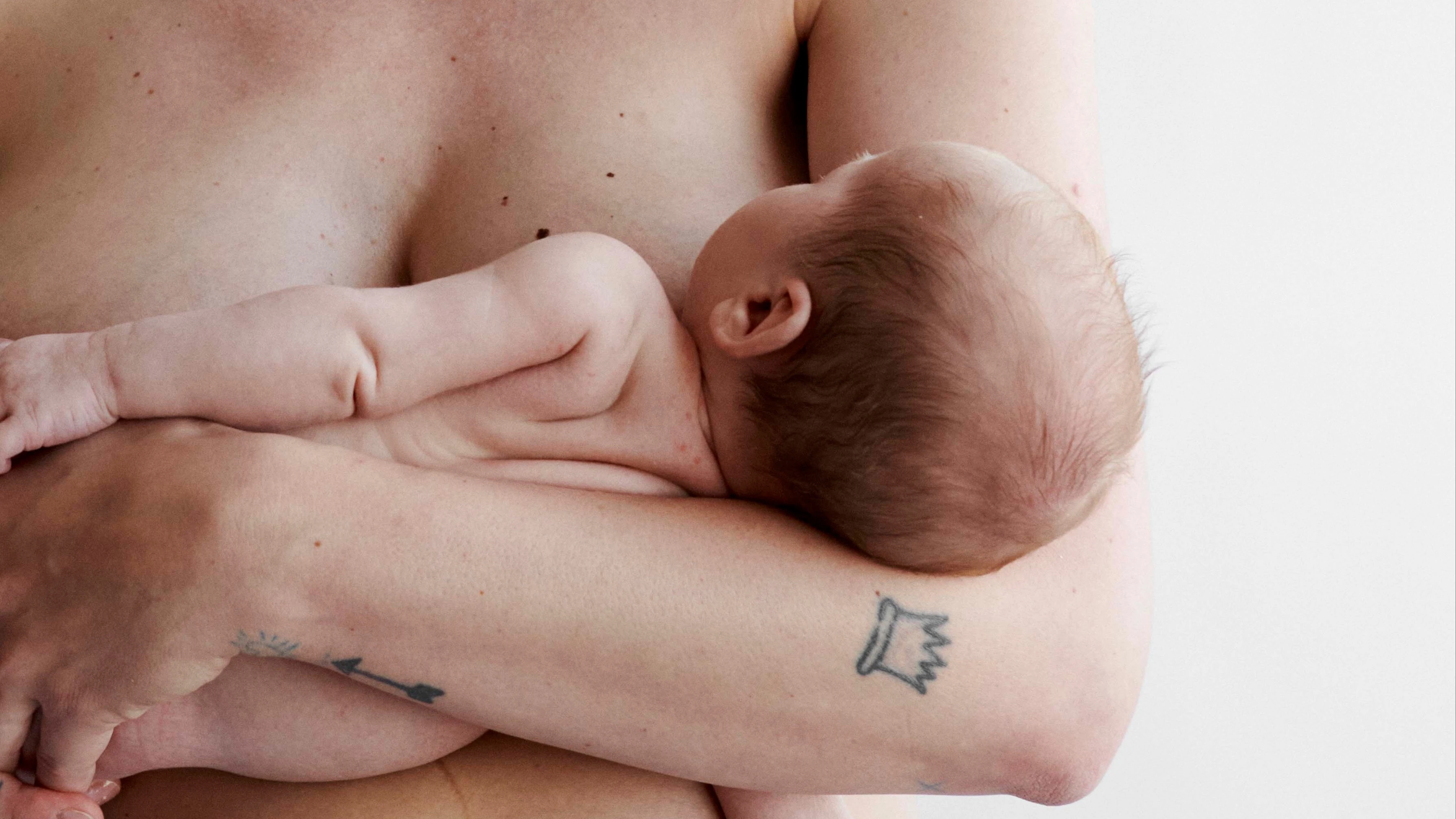 En smilende kvinde med bar overkrop holder blidt en baby i sine arme mod en hvid baggrund.