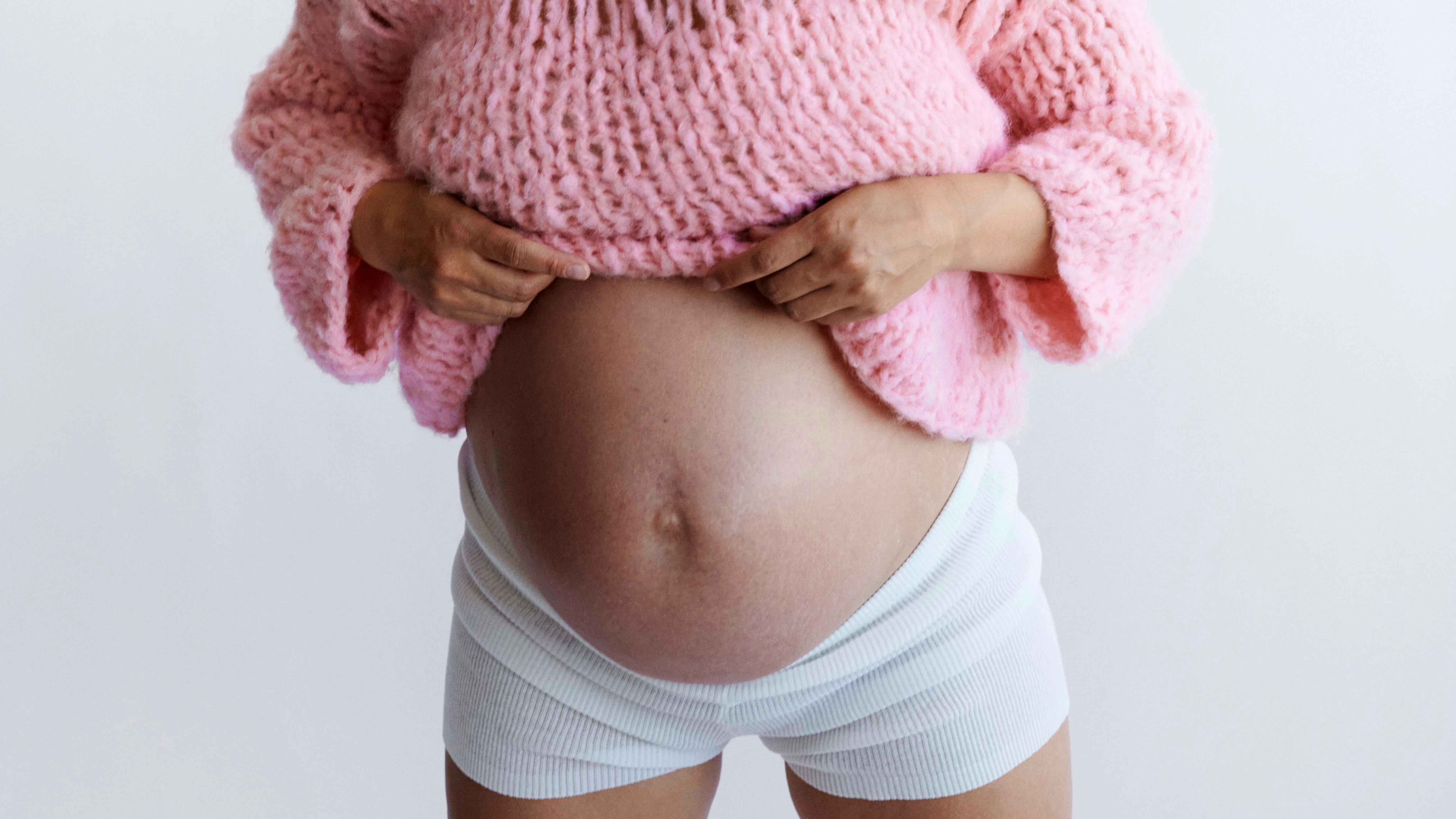 En smilende gravid kvinde iført en lyserød striktrøje og hvide shorts, der viser sin babybule.