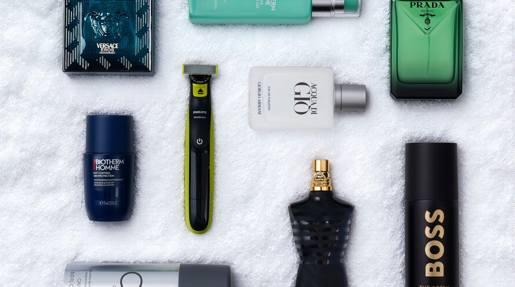 Et fladt layout af pleje- og duftprodukter til mænd: Versace, Biotherm, Philips OneBlade, Prada, Giorgio Armani, Jean Paul Gaultier og Hugo Boss, på et hvidt håndklæde.